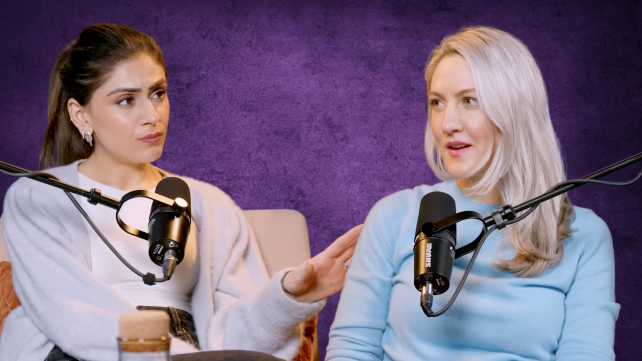 Two women speaking into microphones during a podcast recording, with one woman gently touching the other's shoulder.
