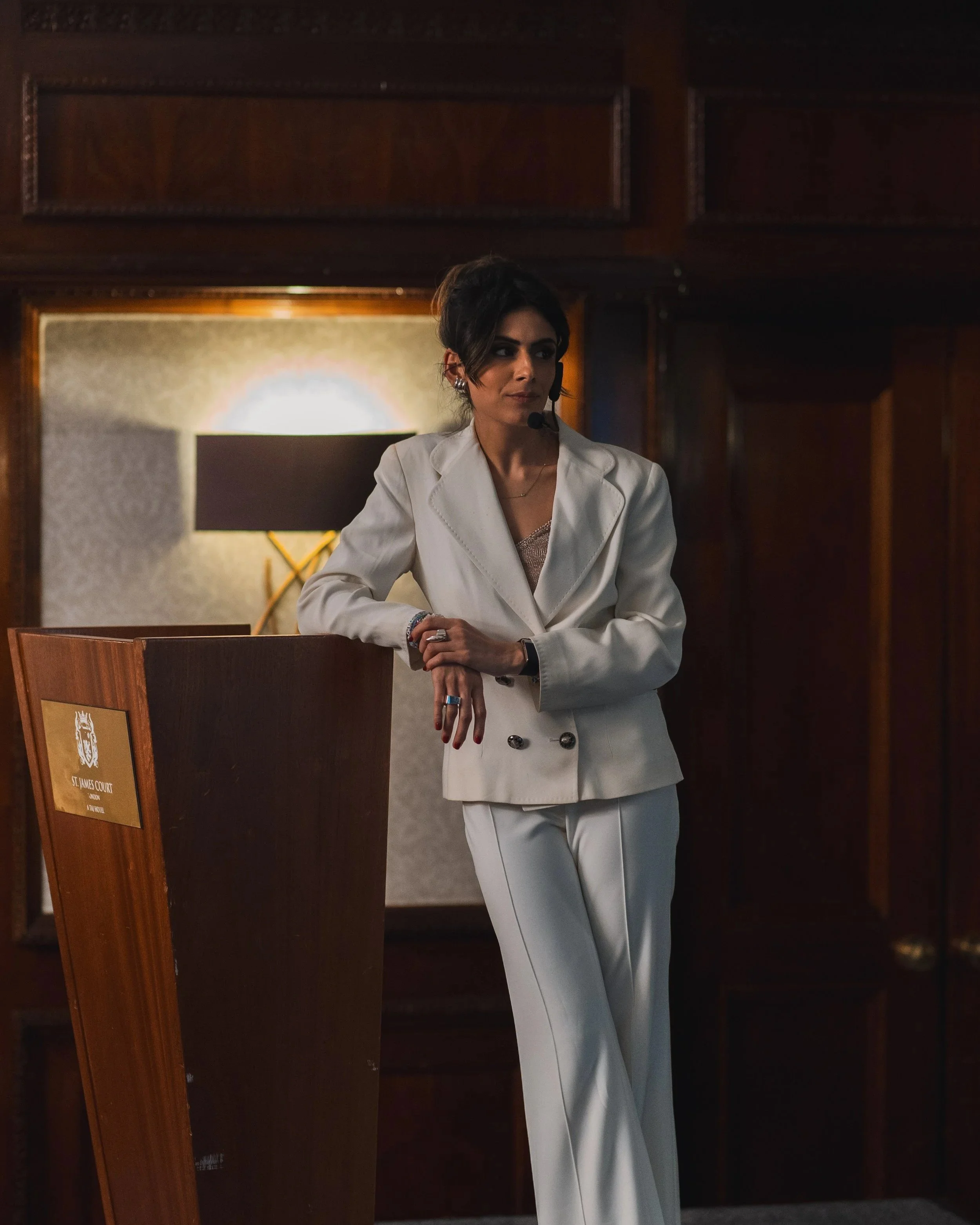 Woman wearing a white business suit with a headset, standing in a wood-paneled room at a podium.