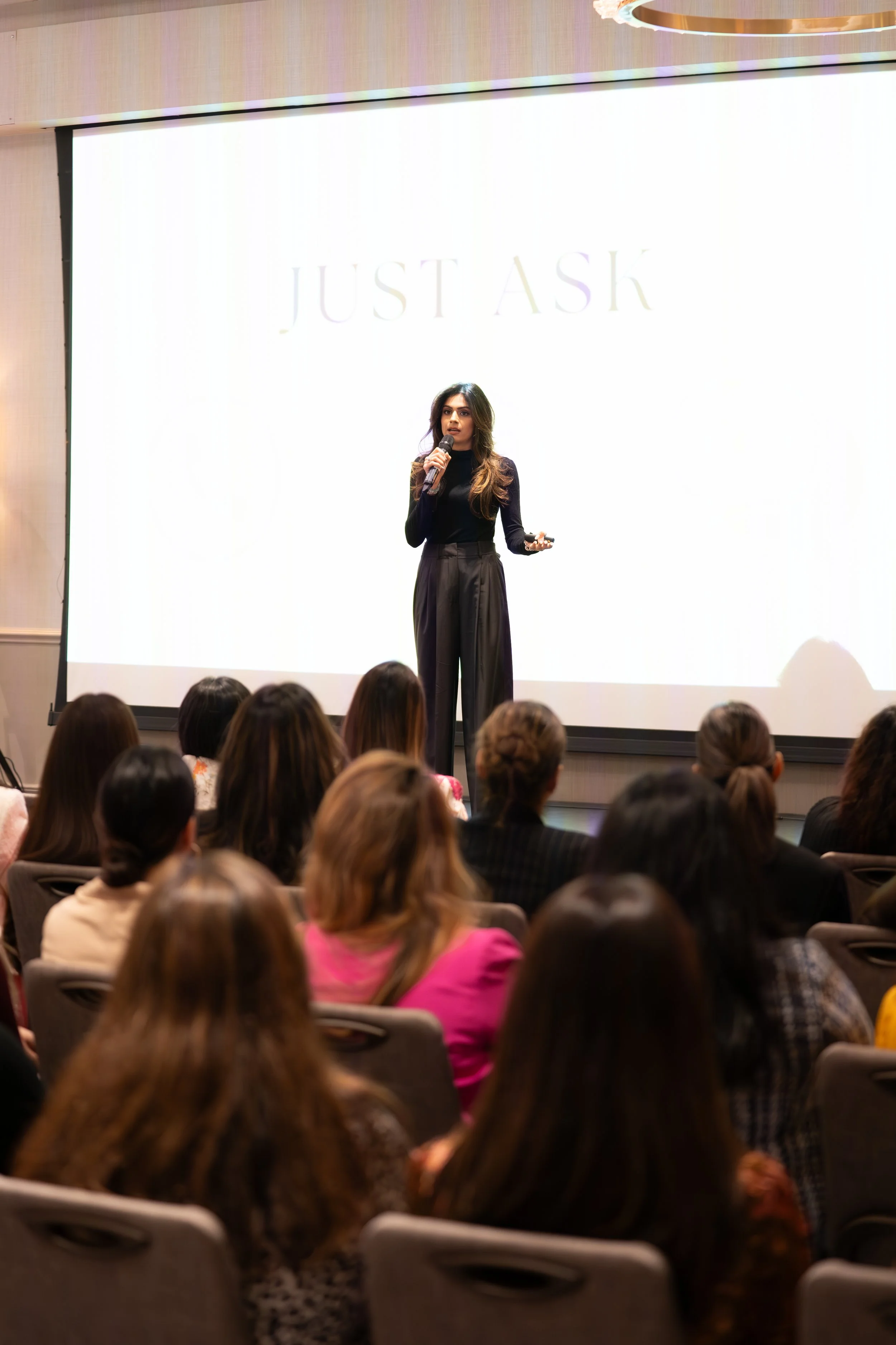 A woman giving a presentation in front of a large, brightly lit screen with the words 'JUST ASK' displayed. She is holding a microphone and standing amidst an audience seated in a conference room.