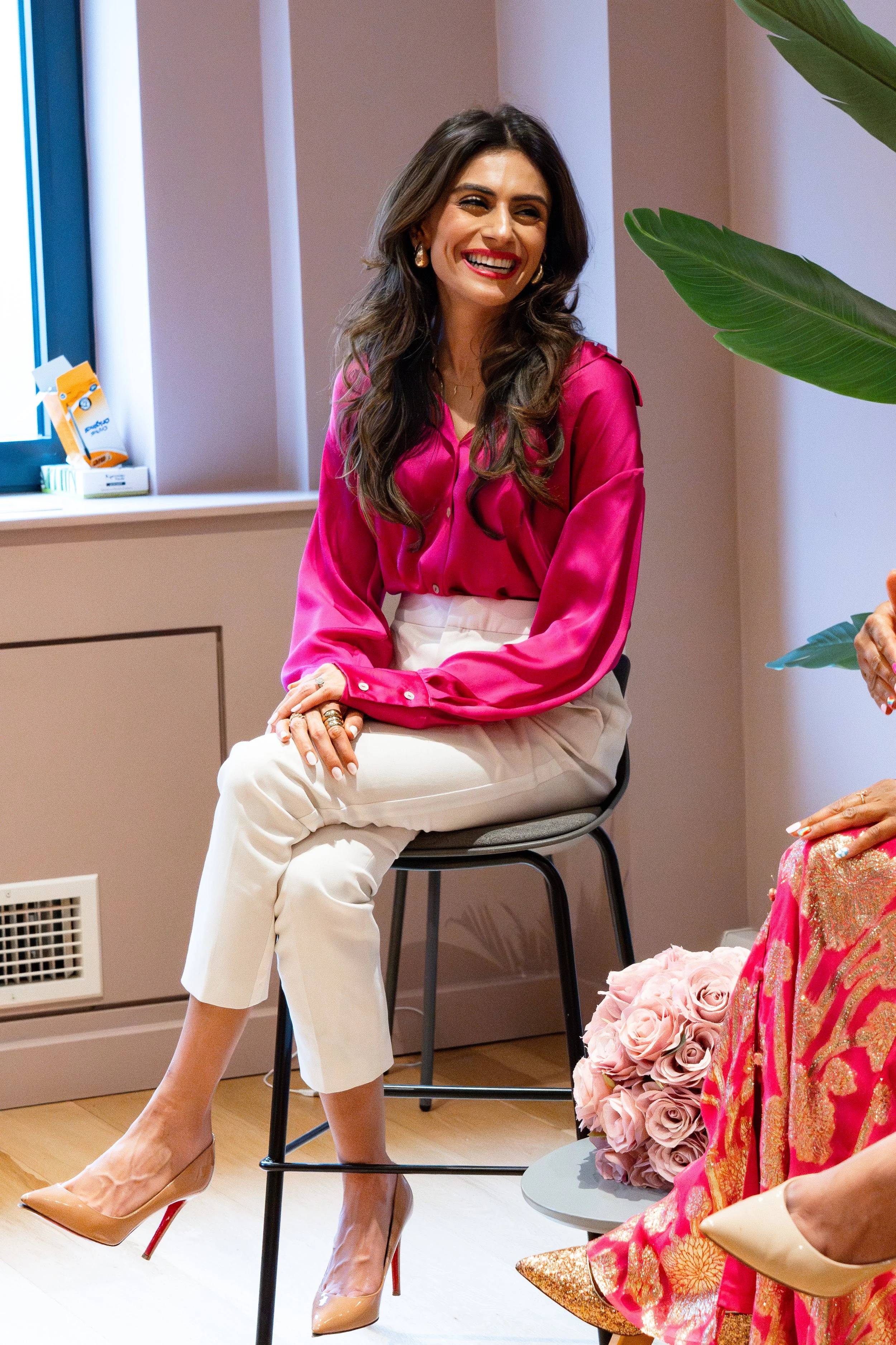A woman with long dark hair wearing a vibrant pink blouse and beige pants, sitting on a gray chair, smiling. There are colorful flowers and large green leaves nearby, with a window and a beige wall in the background.