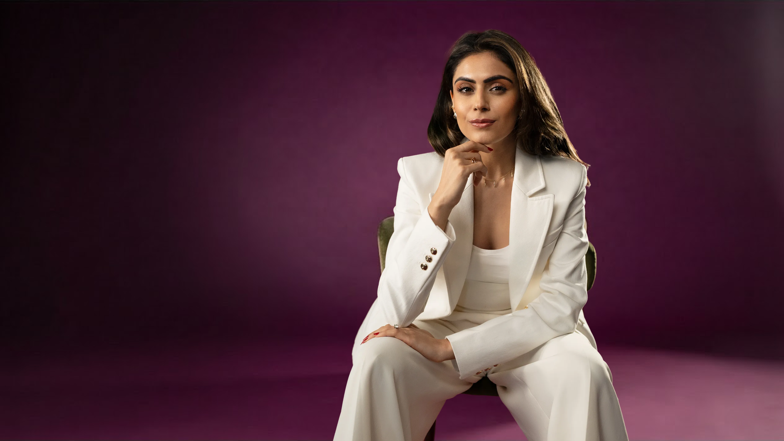 A woman in a white suit sitting on a green chair with a purple gradient background, looking confidently at the camera.