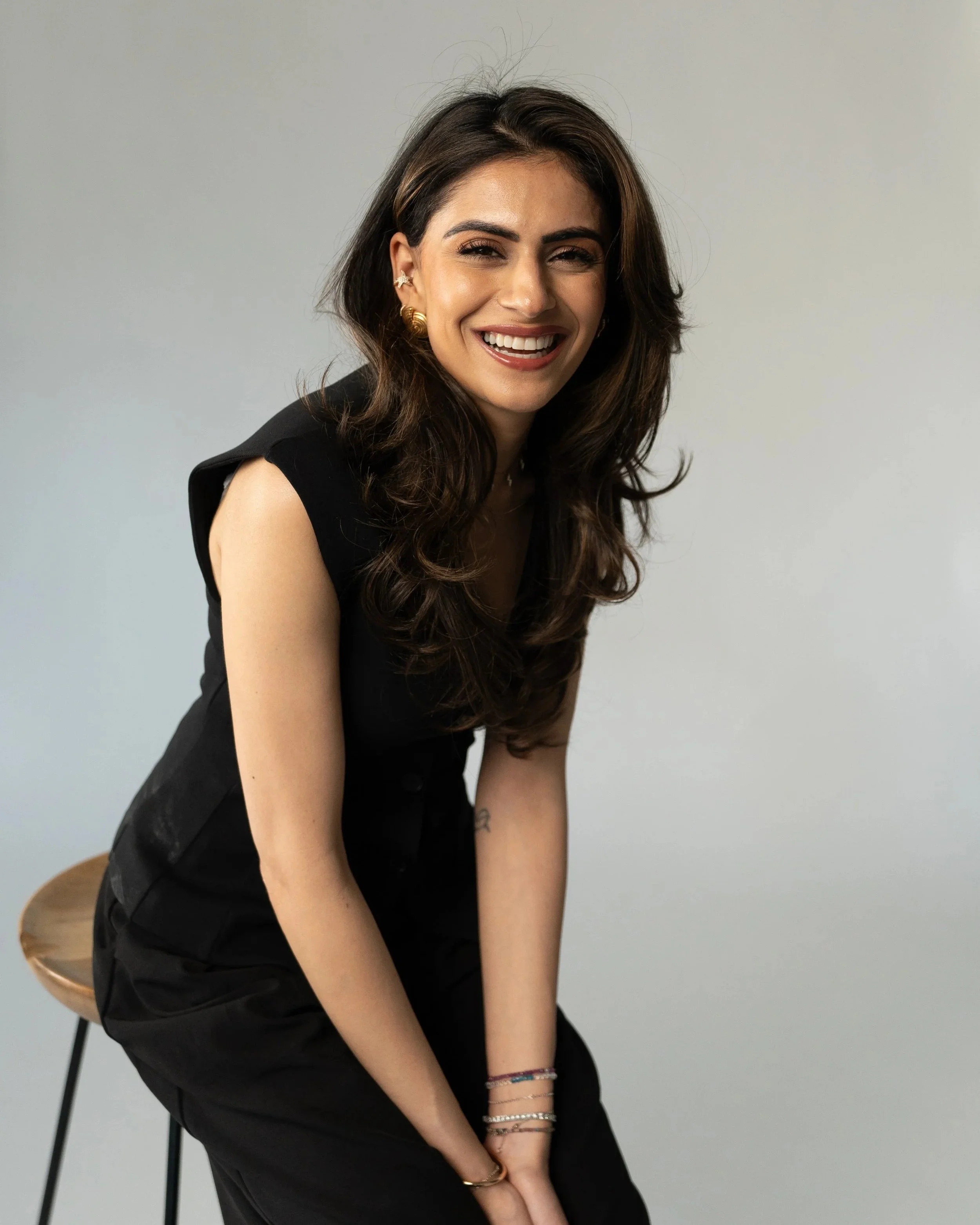 A young woman with long dark hair styled in loose waves, smiling, wearing a black sleeveless top, gold earrings, and multiple bracelets, sitting on a light-colored stool against a plain white background.