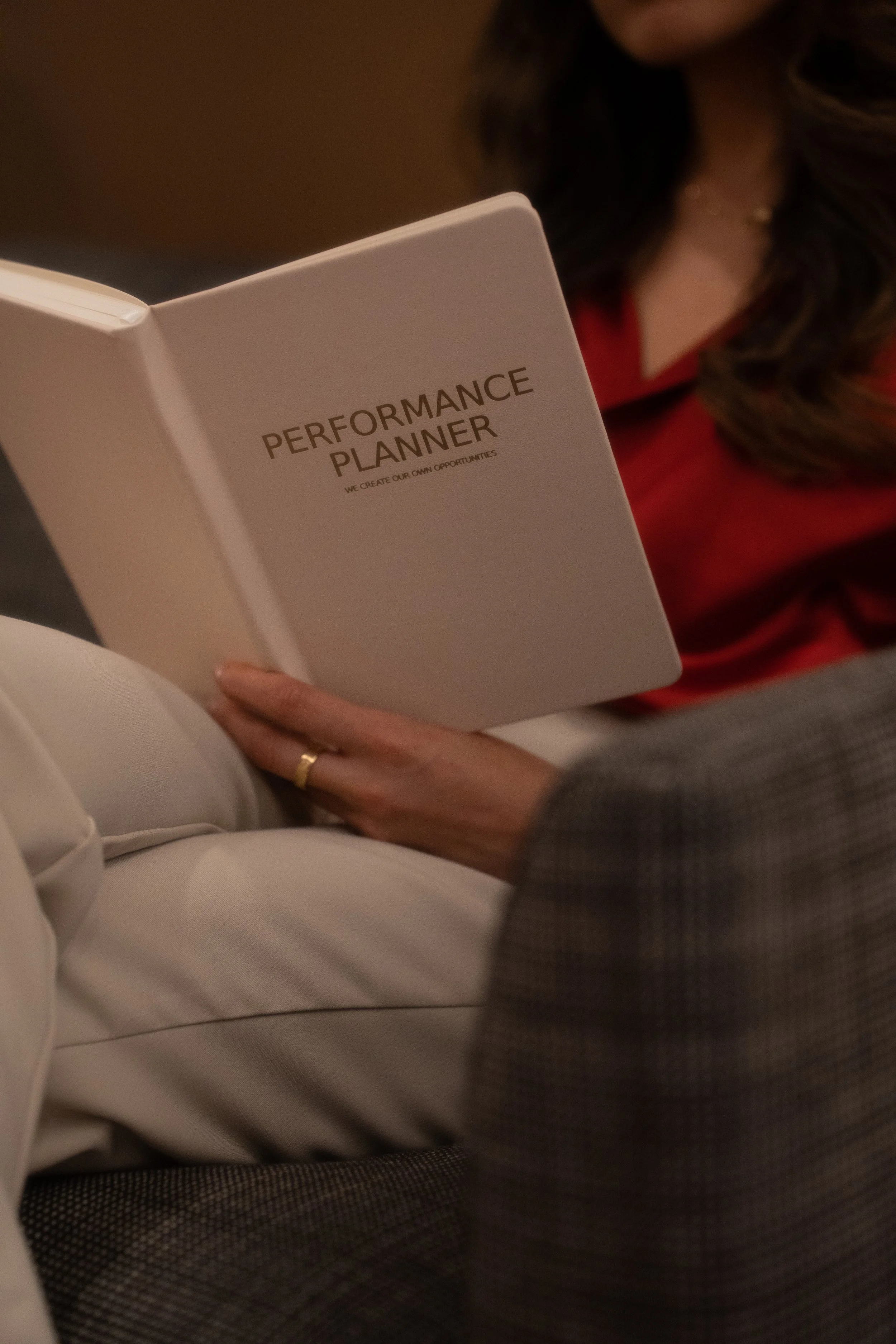 A person in a red top sitting on a white upholstered seat, reading a white book labeled 'Performance Planner' with the subtitle 'We create our own opportunities'. Only part of the person's face and hands are visible.