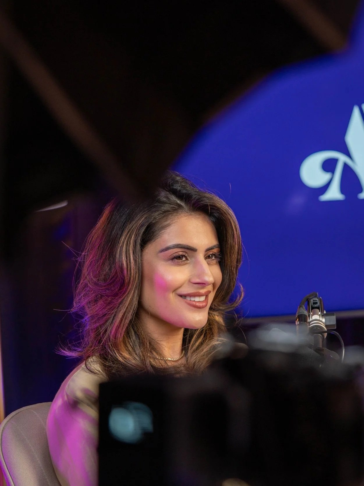 A woman with long wavy brown hair smiling at a press event, in front of a blue background with white logo. The image is partially obscured by a black object and a camera lens in the foreground.