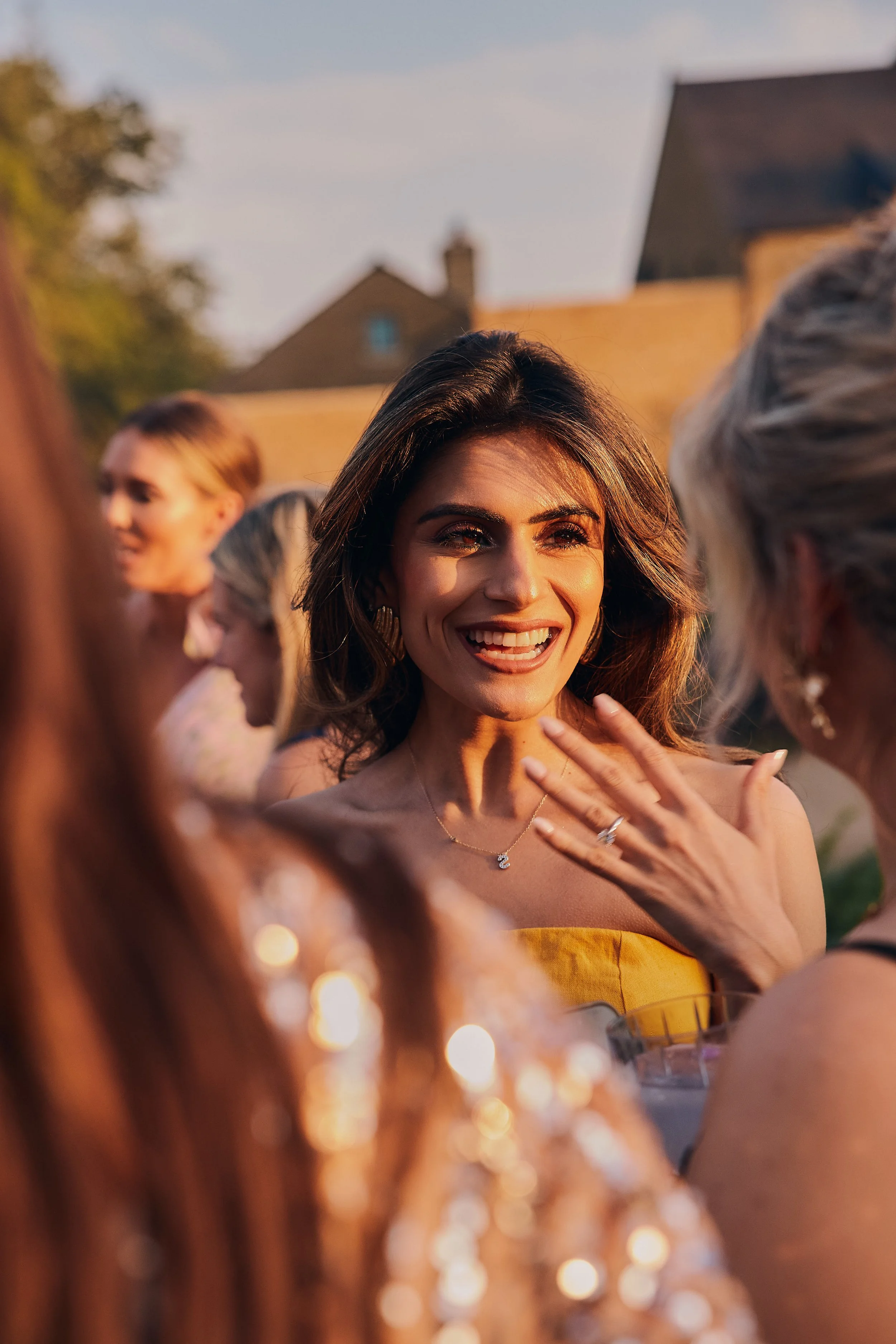 A woman smiling and talking among friends at an outdoor gathering, with warm sunset lighting and blurred background of houses and trees.