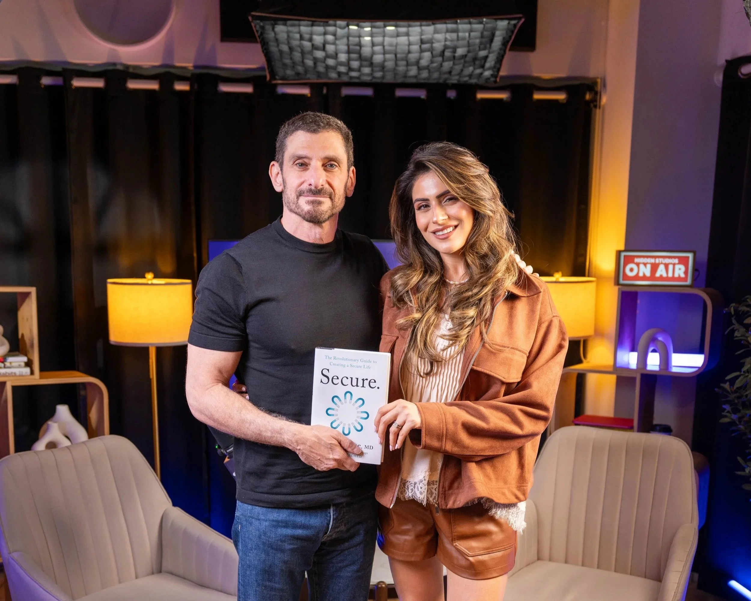 A man and a woman stand closely together, smiling for the camera in a room with dark curtains, yellow lamps, a sign that says 'ON AIR,' and comfortable chairs.