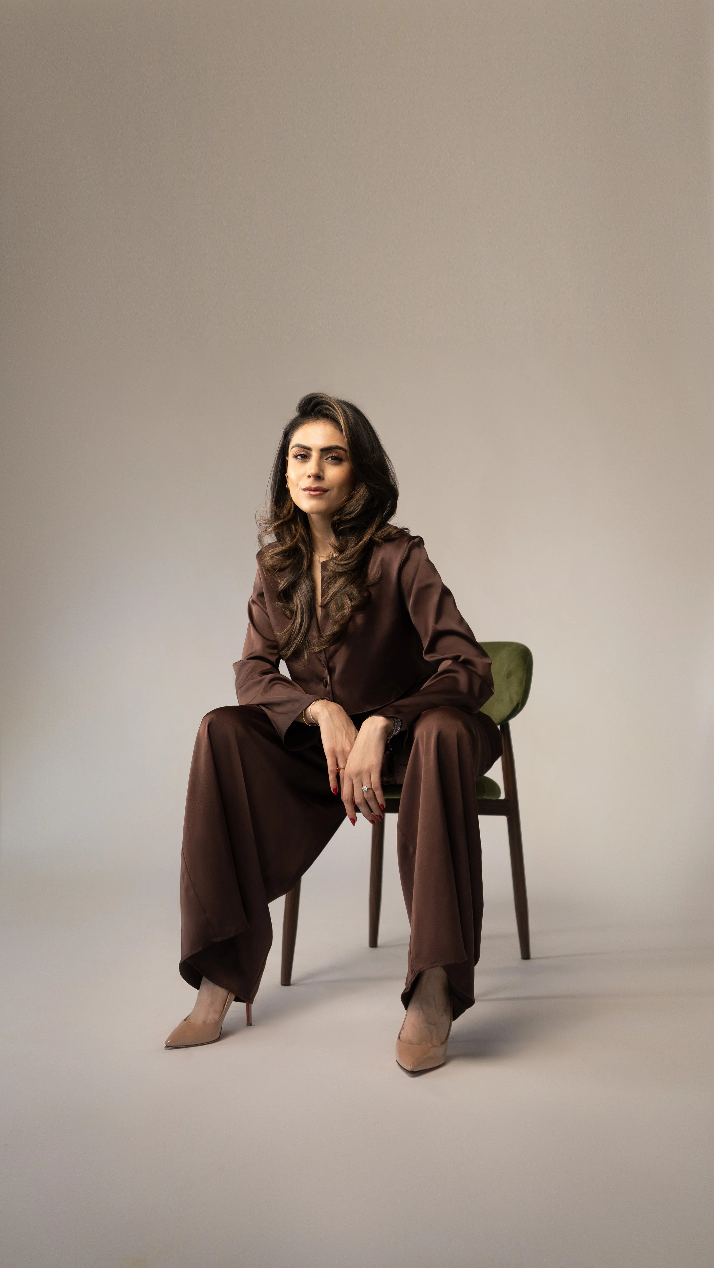 A woman with long dark hair sitting on a green chair, wearing a brown satin pantsuit and beige high heels, against a plain neutral background.
