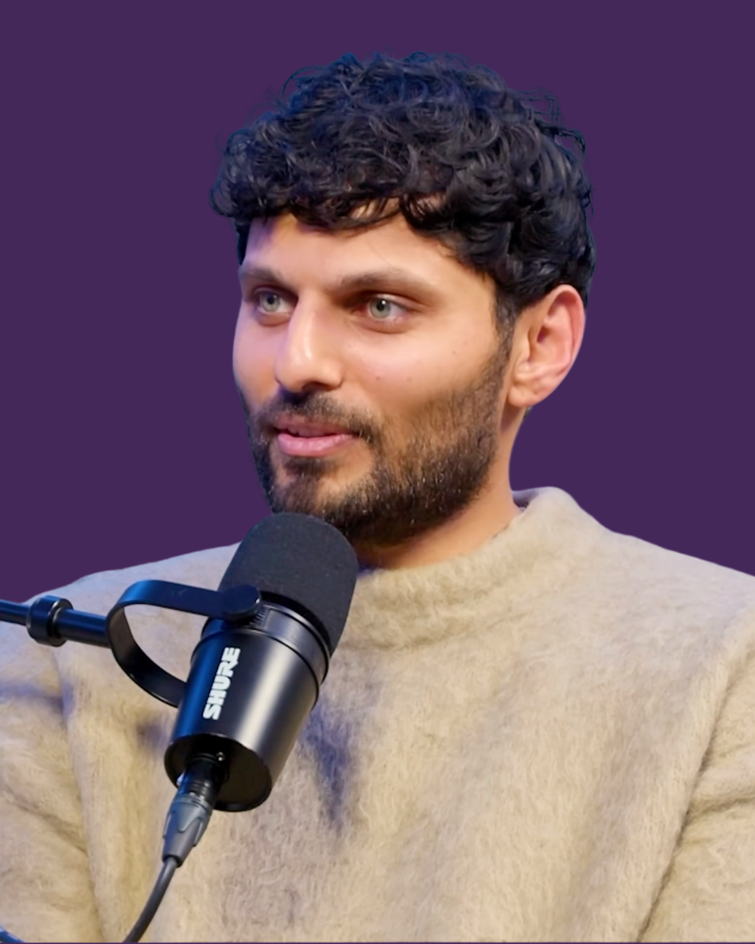A man with curly dark hair and a beard speaking into a black Shure microphone against a purple background.
