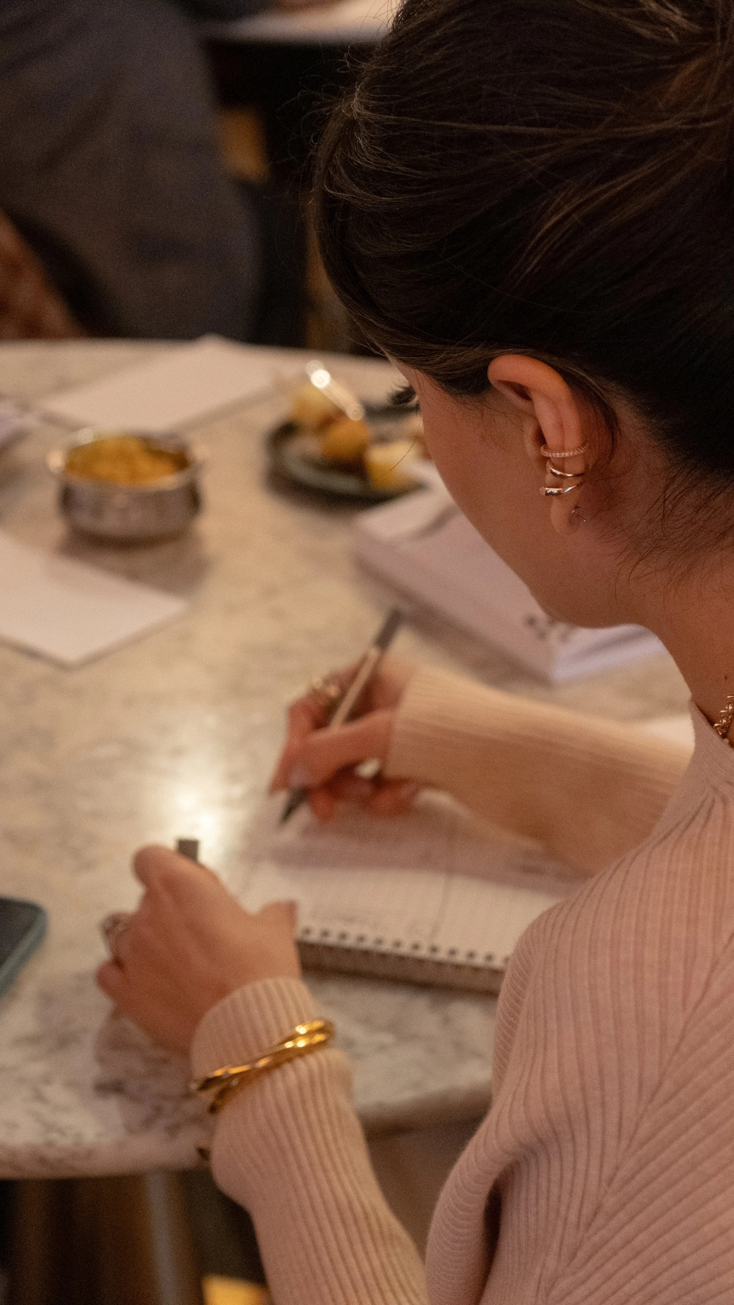A woman with brown hair, earrings, and a watch writing in a notepad at a table with food and drinks.