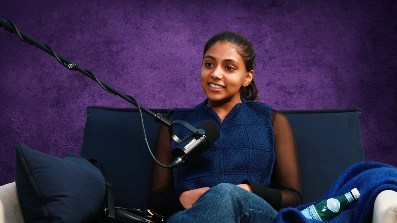 Young woman sitting on a dark blue couch with a purple textured wall background, speaking into a microphone.
