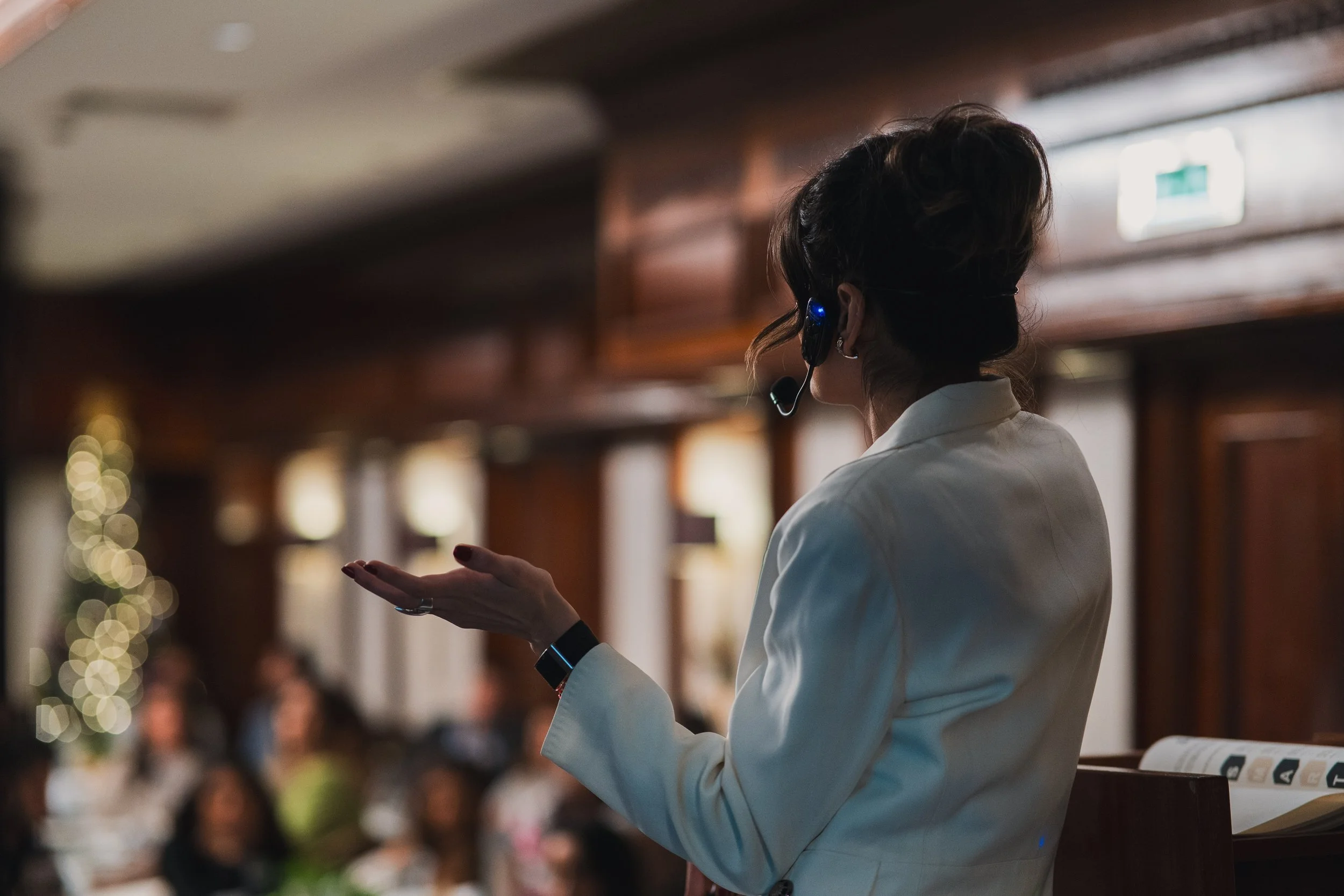 A woman in a white blazer speaking at an indoor event, wearing a headset microphone, with a blurred audience and warm lighting in the background.
