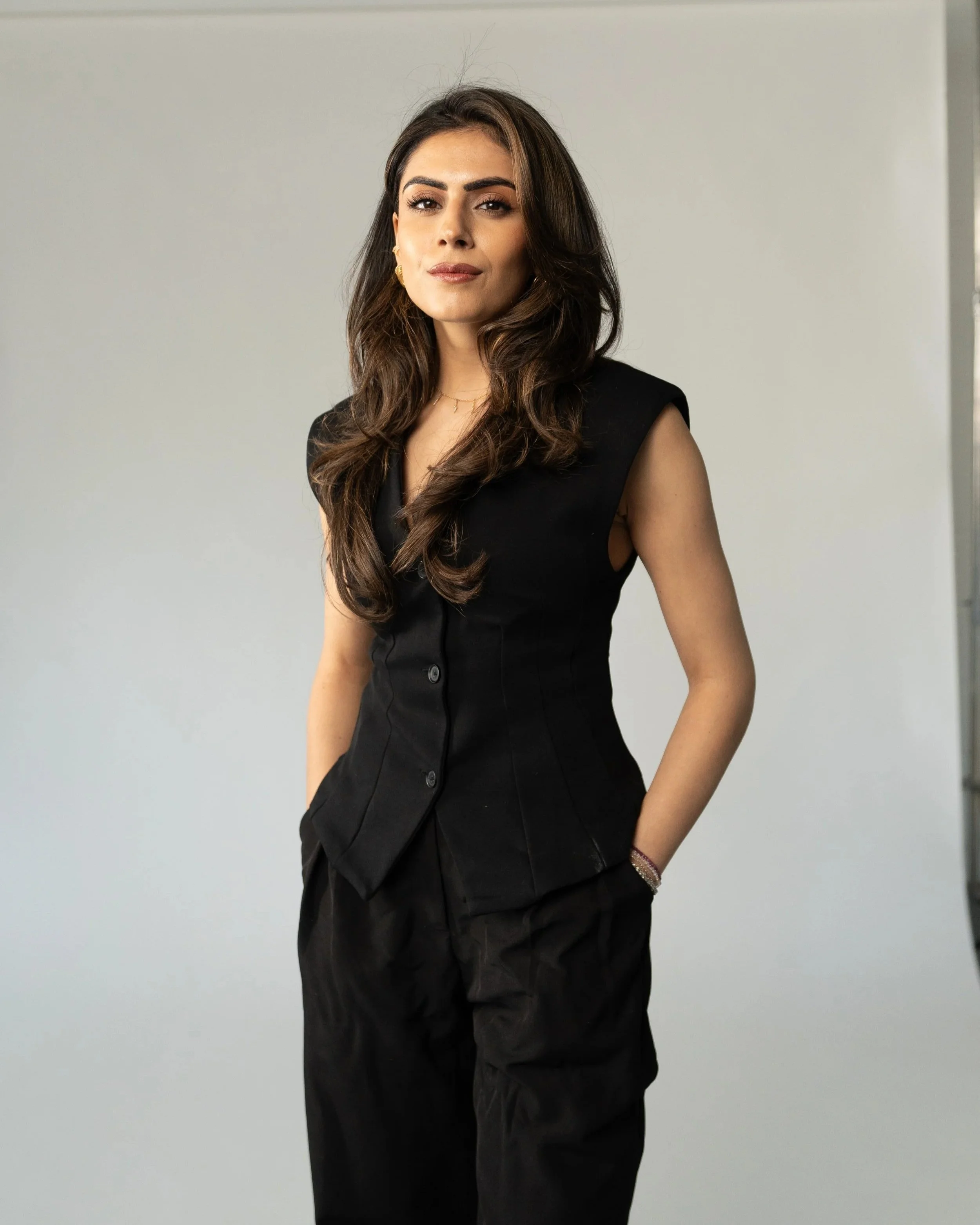 A woman with long brown hair, wearing a black sleeveless top and earrings, posing against a plain light gray background.