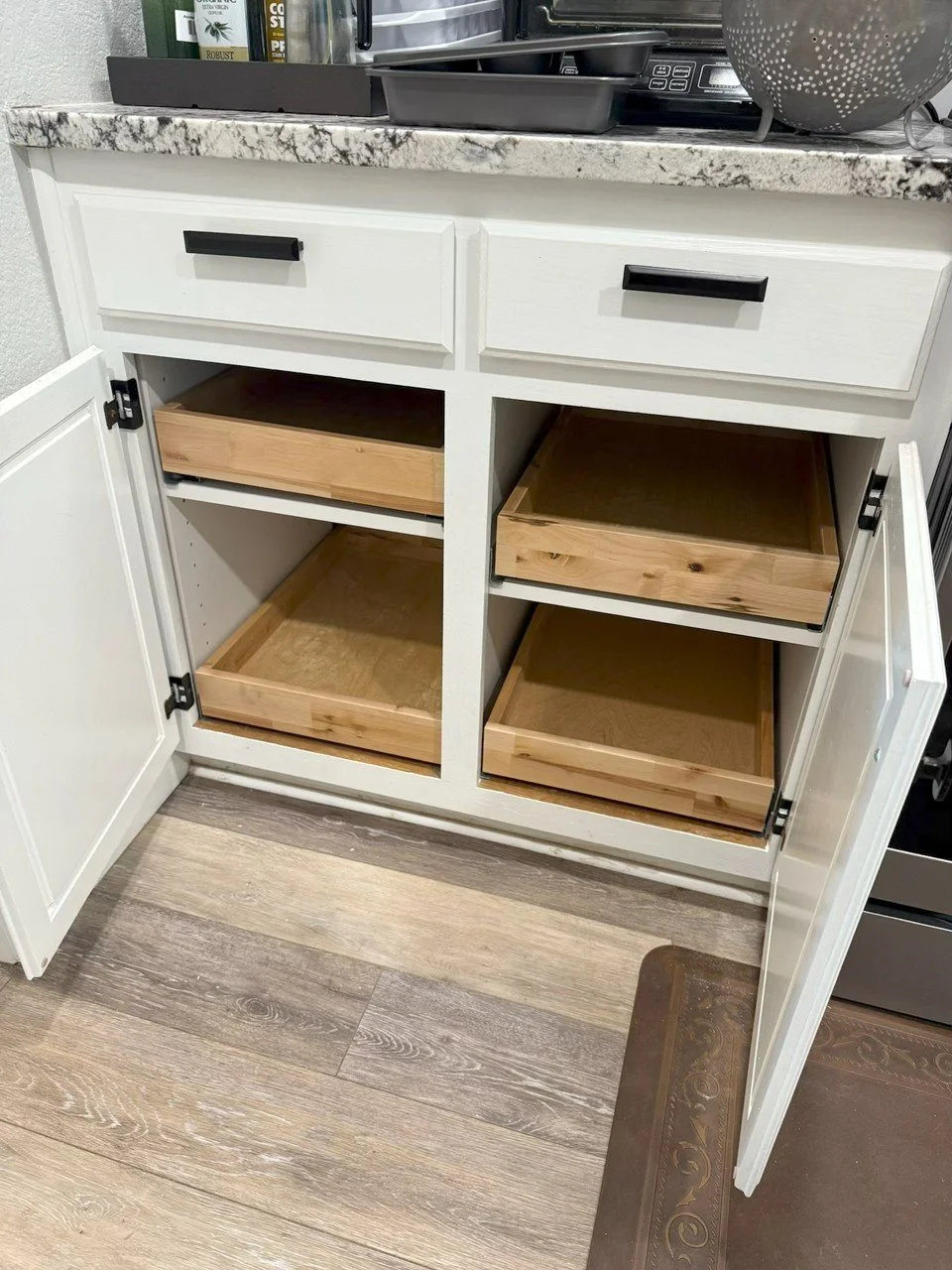 Open kitchen cabinet with two wooden trays inside, situated beneath a granite countertop.