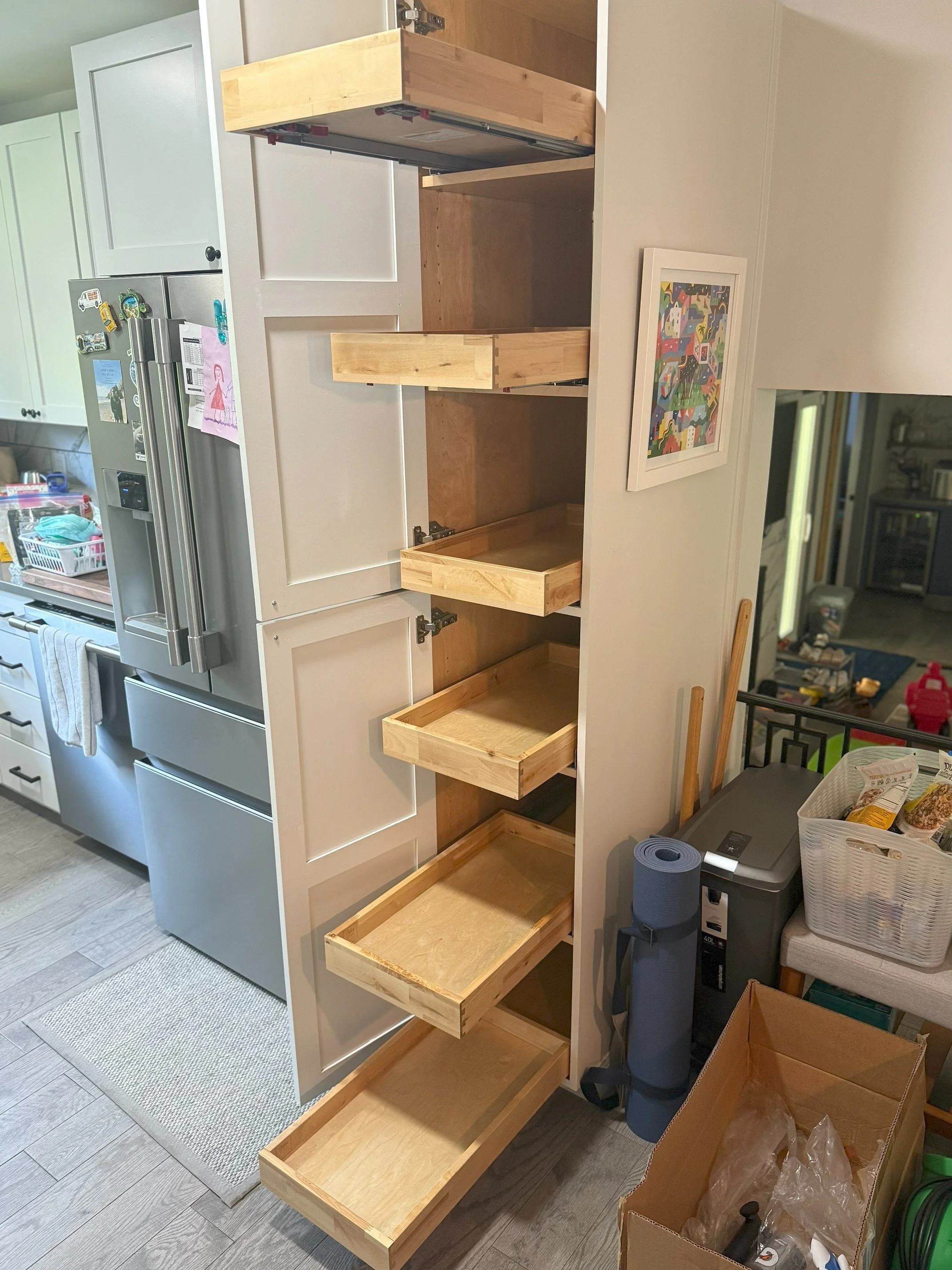 An unfinished set of six wooden drawers installed into a white kitchen cabinet. The drawers are open and appear to be in the process of being assembled or customized. The corner of a kitchen is visible in the background, with a stainless steel refrigerator, a white towel hanging, and some magnets and papers on the fridge. To the right, there's a colorful artwork on the wall, and a glimpse of a living room area with furniture, a red toy, and various items.