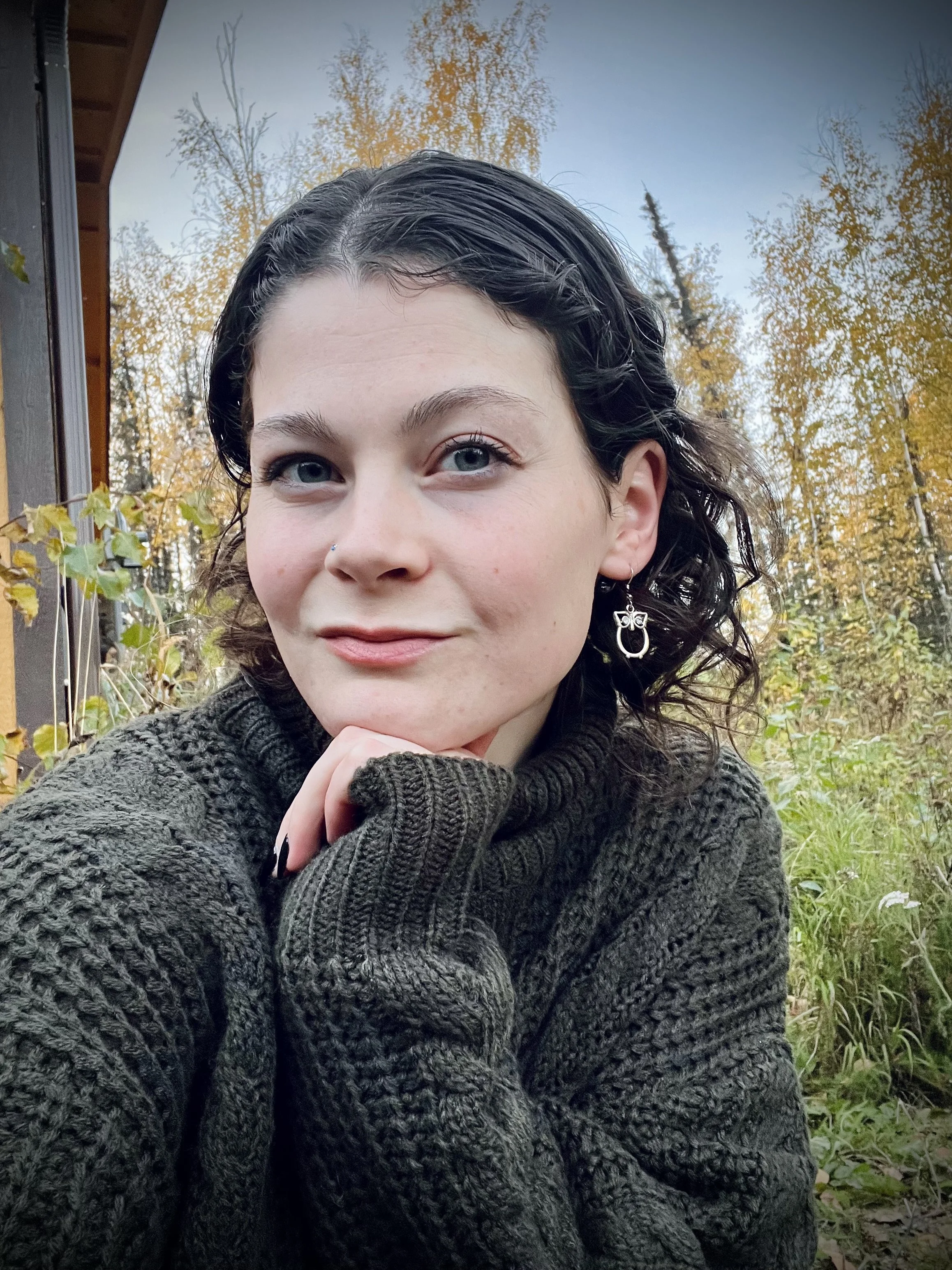 A woman with dark, curly hair and blue eyes sitting outdoors among trees with autumn foliage, wearing a dark textured sweater and earrings, looking at the camera.