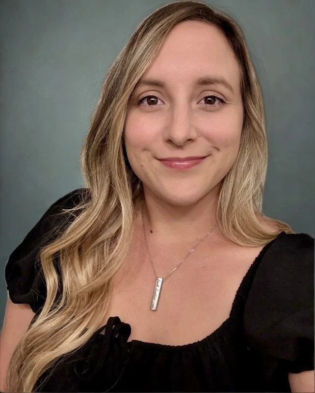 A woman with long wavy blonde hair smiling at the camera, wearing a black top with puffed sleeves and a silver pendant necklace with engraved design, standing against a plain gray-green background.