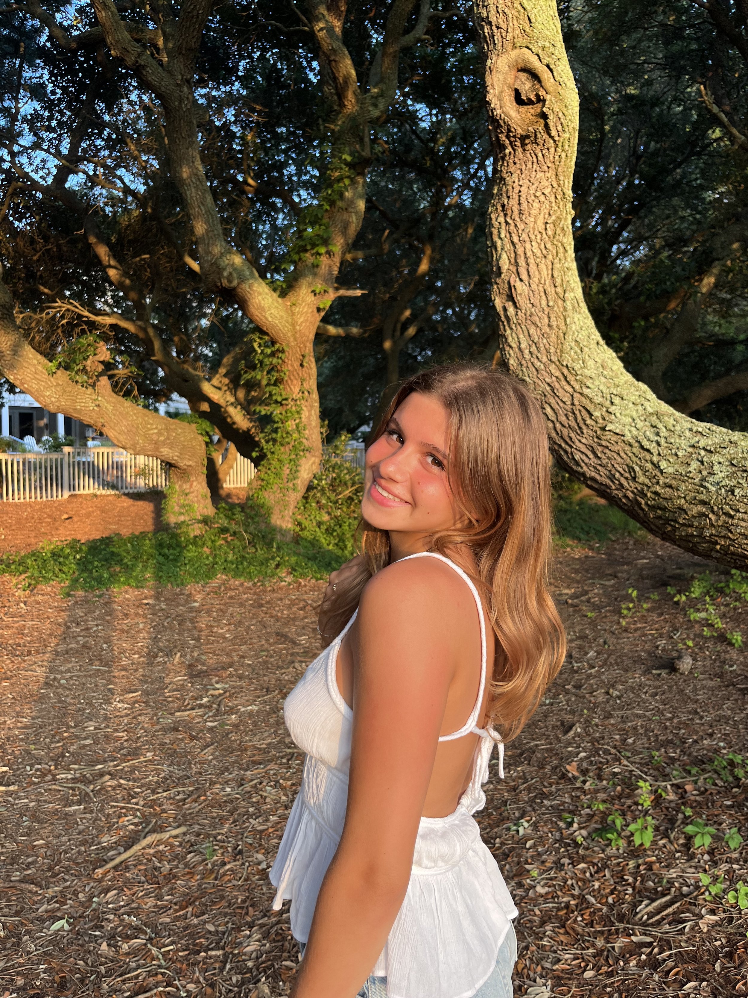 A young woman with long, wavy, light brown hair and a bright smile is standing outdoors next to a large tree with textured bark. She's wearing a white sleeveless top and light-colored bottoms. The background includes the tree, some greenery, and a white fence, all illuminated by warm, natural sunlight.
