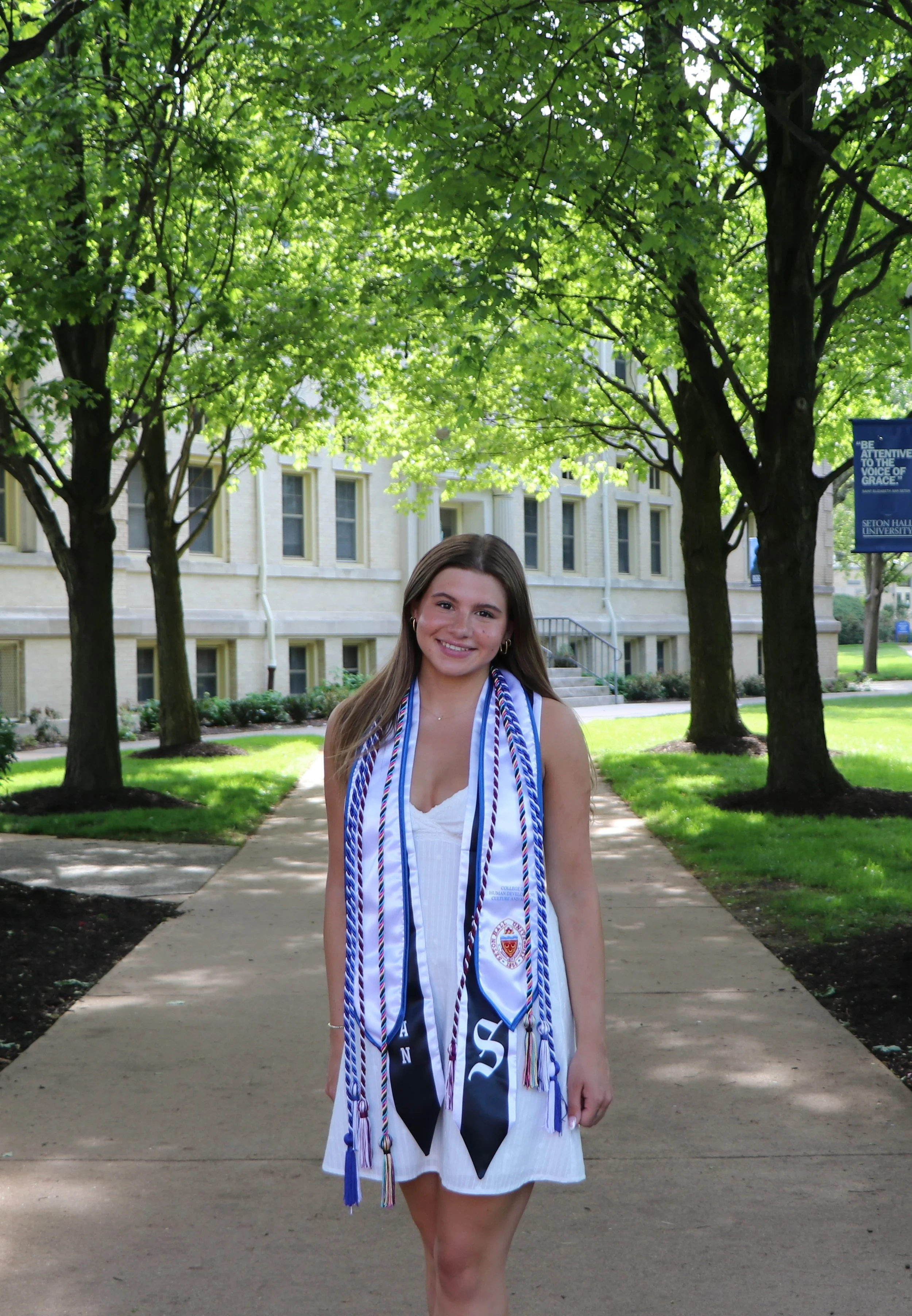A young woman in a white dress wearing multiple graduation cords and medals, standing outdoors on a sidewalk with green trees and a building in the background.