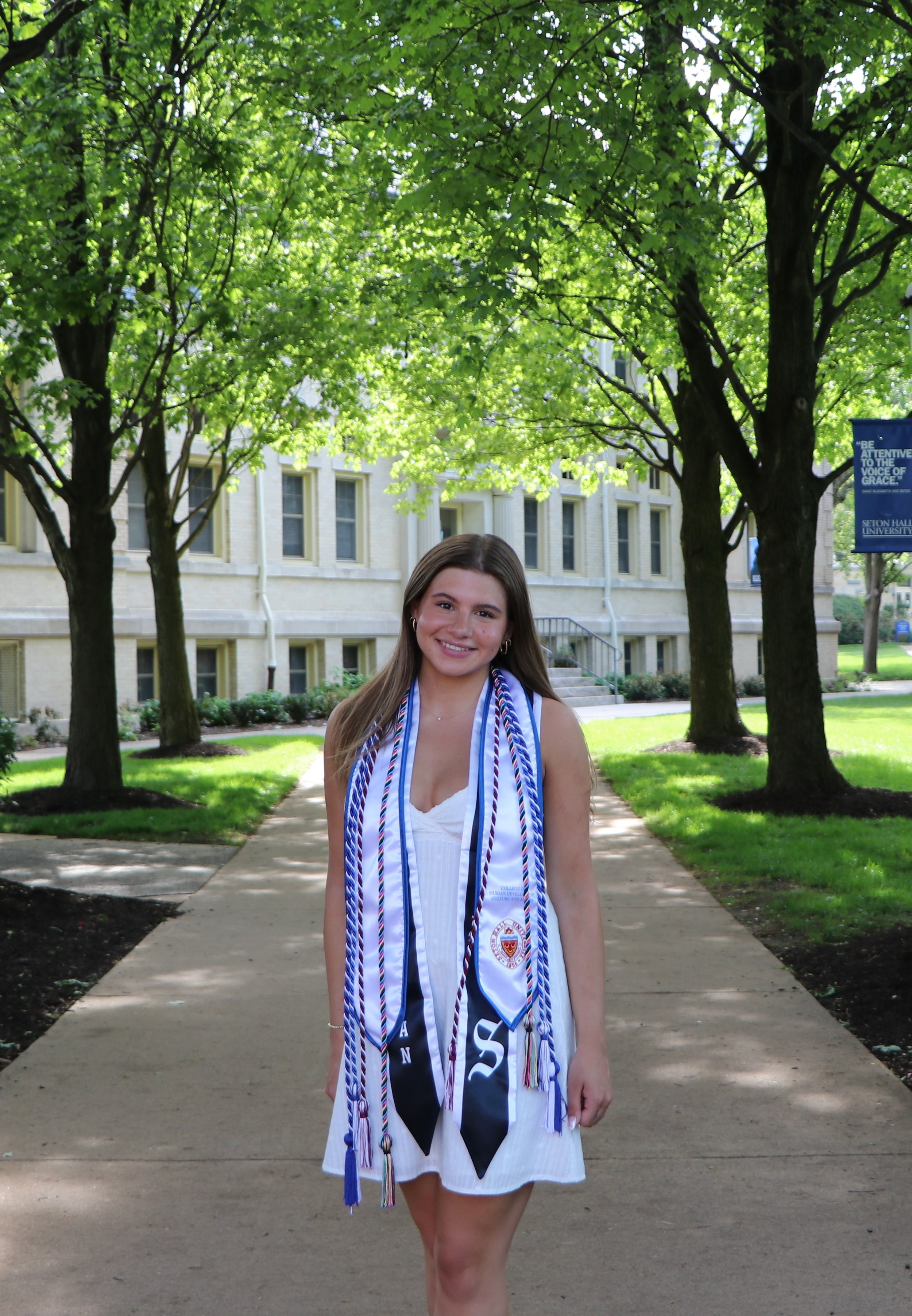 A young woman in a white dress and graduation gown with numerous blue and white honor cords around her neck, standing on a sidewalk outdoors with trees and a university building in the background.