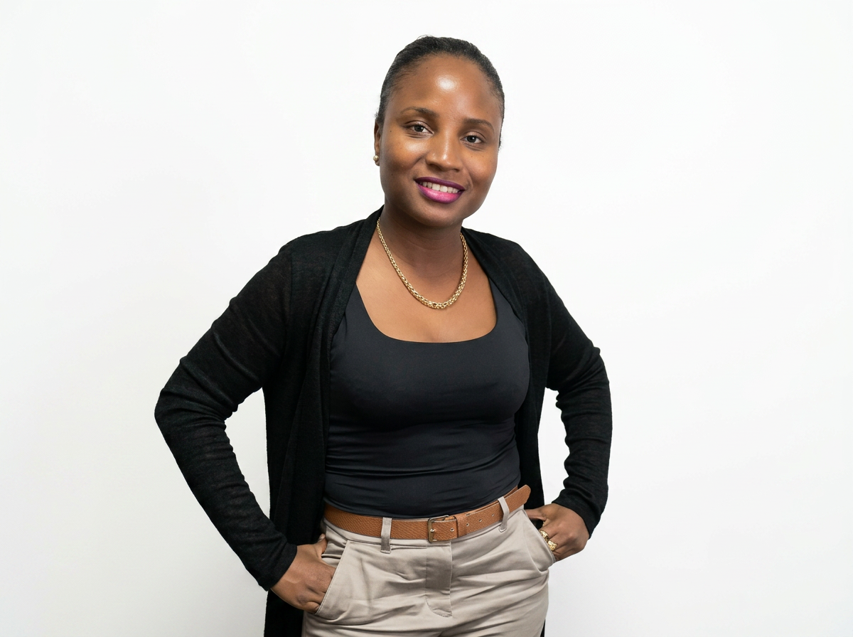 Une femme souriante posant avec les mains sur les hanches, portant une chemise noire, un cardigan noir et un pantalon beige, avec un fond blanc.