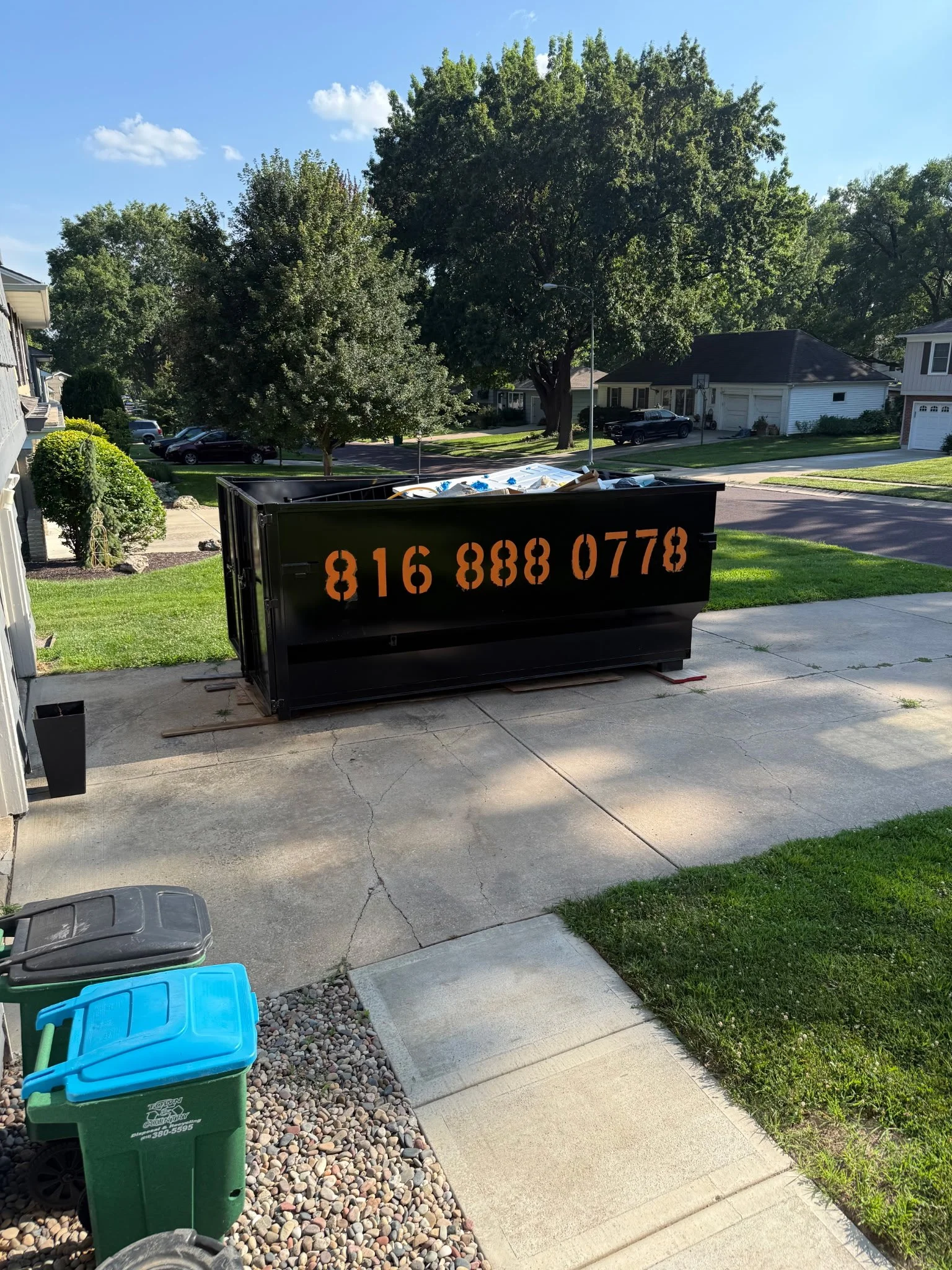 Residential neighborhood on a sunny day with a black dumpster filled with debris in the driveway, green grass, trees, parked cars, and a blue sky with a few clouds.