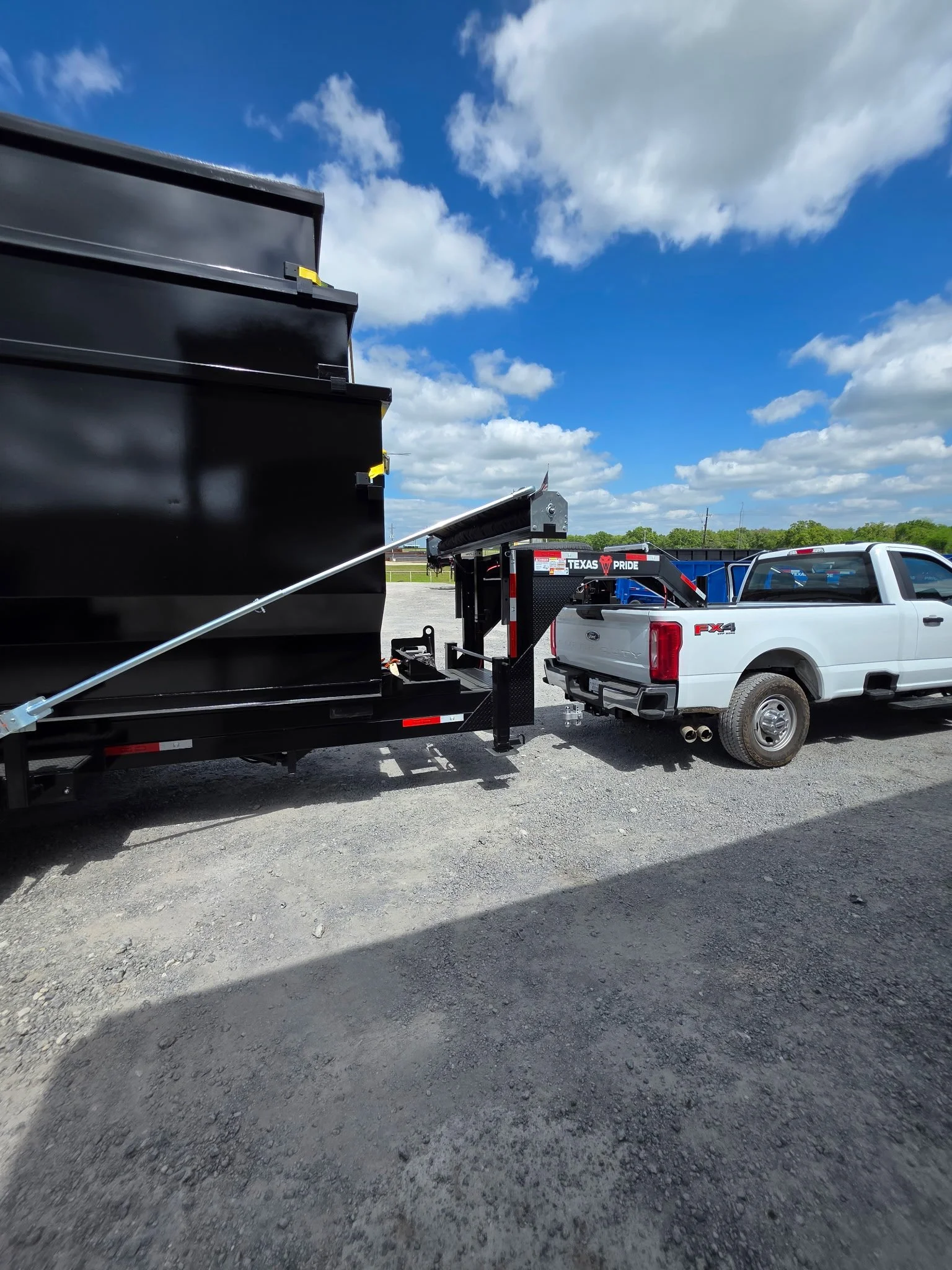 A white pickup truck with a trailer hitch connected to a black trailer. The trailer is attached to a tow vehicle with 'Texas Pride' branding. The photo shows a partly cloudy sky over a gravel lot, with some trees and utility poles in the distance.