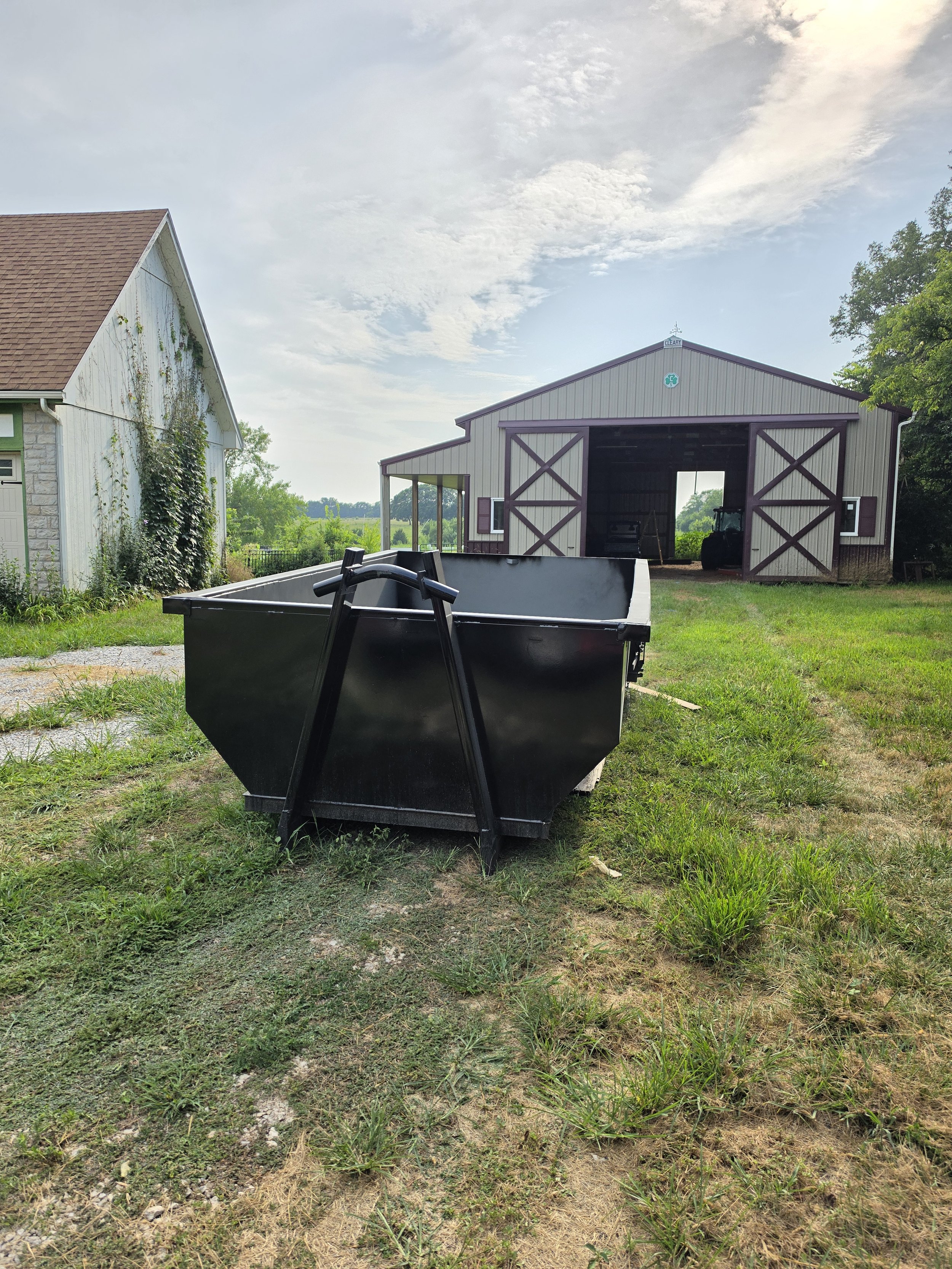 A black dumpster on a grassy patch of land with a large barn in the background.