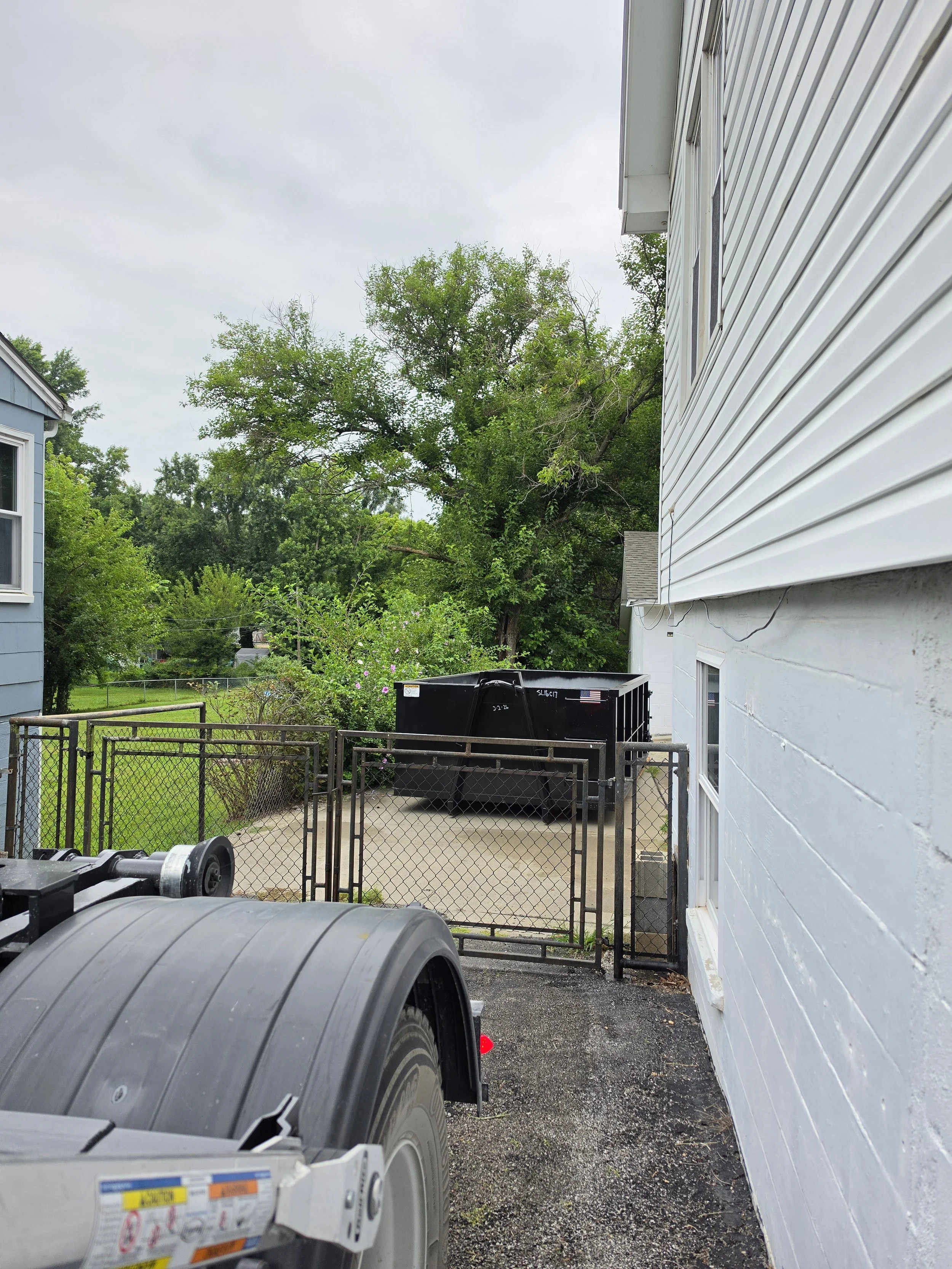 View of a backyard with green trees, a chain-link fenced area, two large black trash containers, and a white house with vinyl siding on a cloudy day.