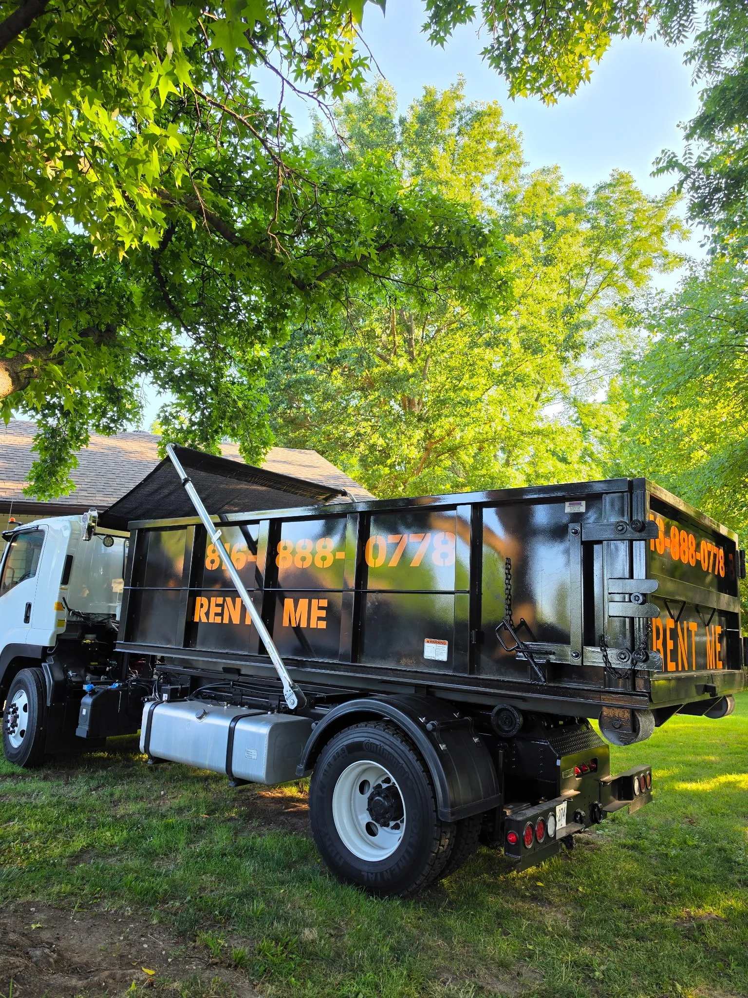 Side view of a black roll-off dumpster truck with orange text on the side, parked on grass under green trees on a sunny day.
