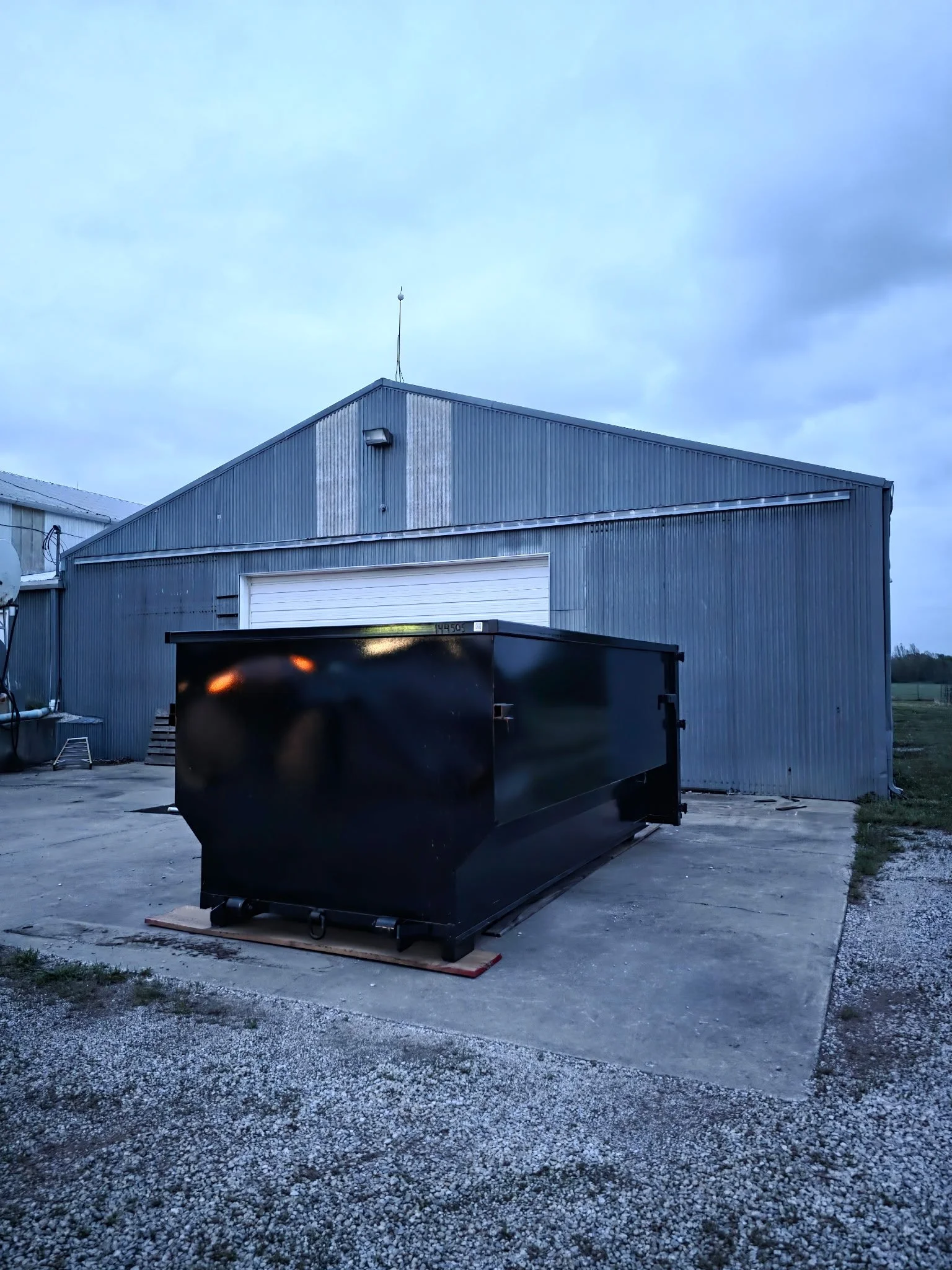 A large metal industrial building with a closed garage door, situated on a concrete pad, with a black dumpster in the foreground and cloudy sky above.