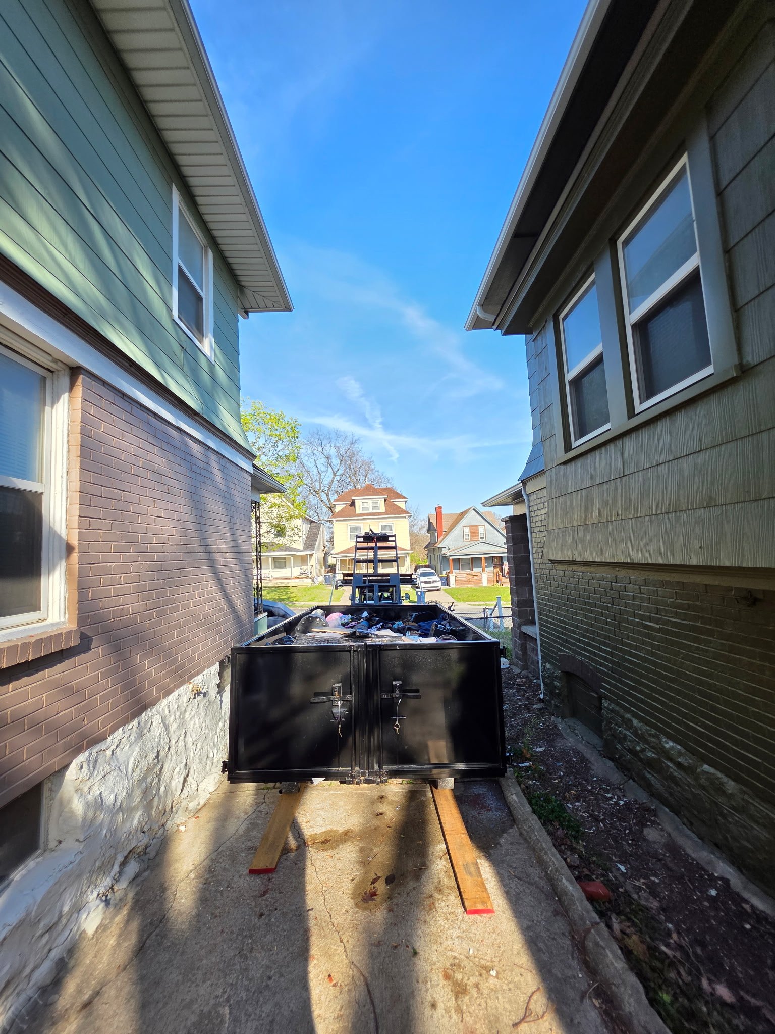 View of a narrow alleyway between two houses, with construction materials and a trailer filled with debris at the end, under a clear blue sky.