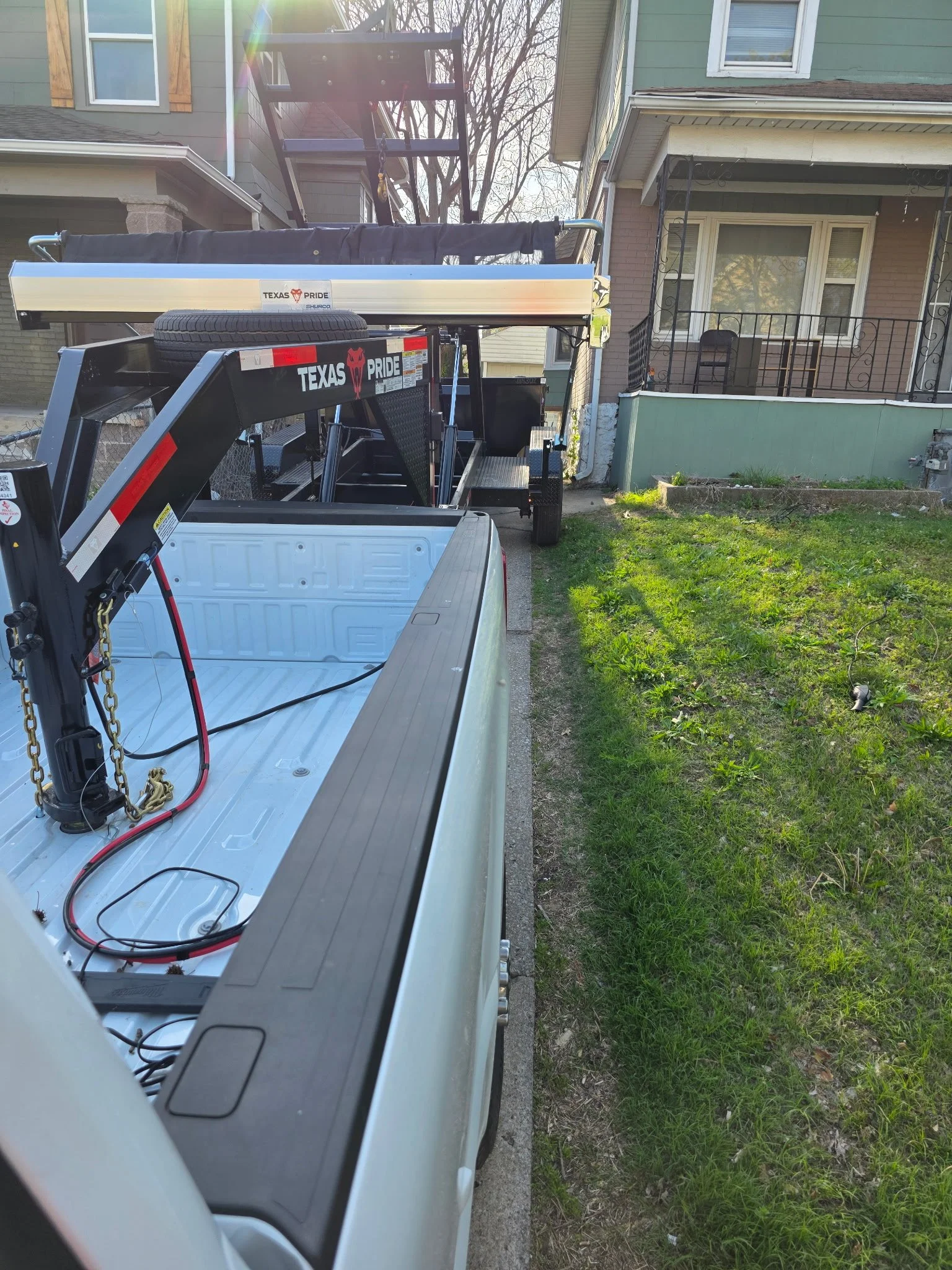 A flatbed truck with a trailer attached parked in front of a house on a driveway, with houses, trees, and grass visible in the background.