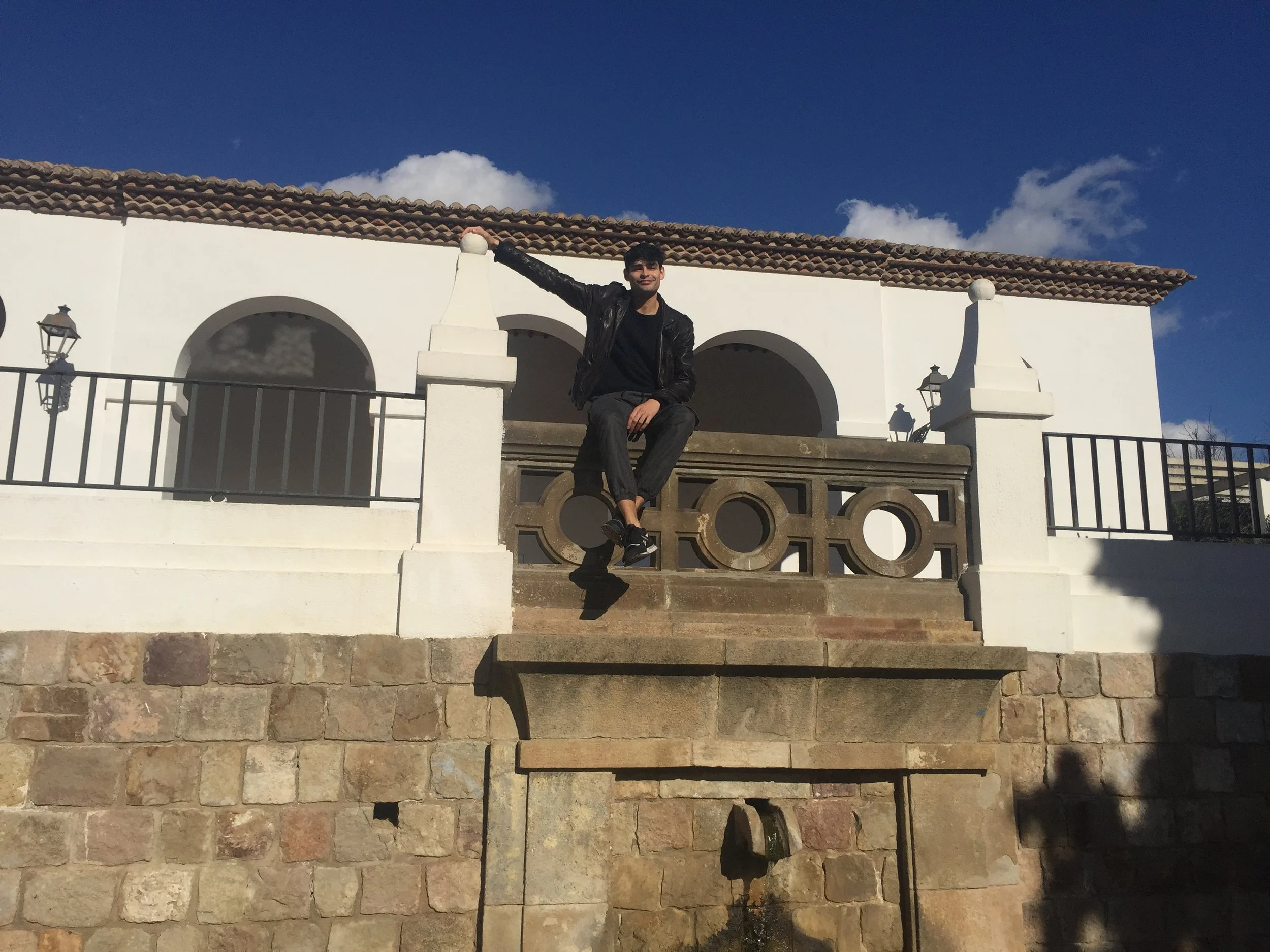 A young man with dark hair, wearing a black leather jacket and black pants, sitting on a stone staircase and leaning against a white railing, with a white building behind him and a blue sky with some clouds overhead. Barcelona