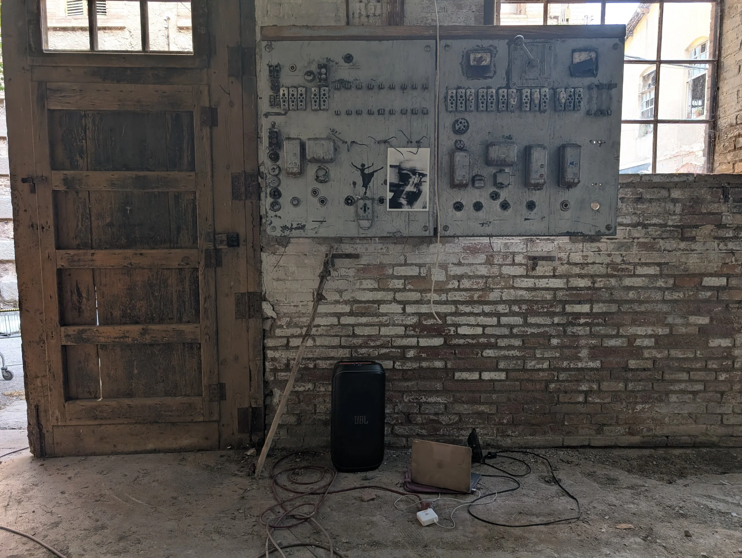 Interior view of a rustic brick and wooden wall with a panel of electrical control switches and circuit breakers. There are wires and electronic devices on the ground, and a window with a view of buildings outside.
