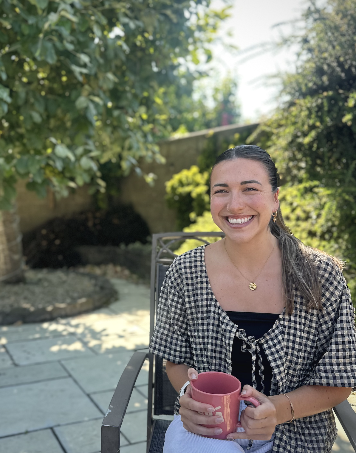 A woman smiling and holding a pink mug while sitting outdoors in a backyard with trees and plants.