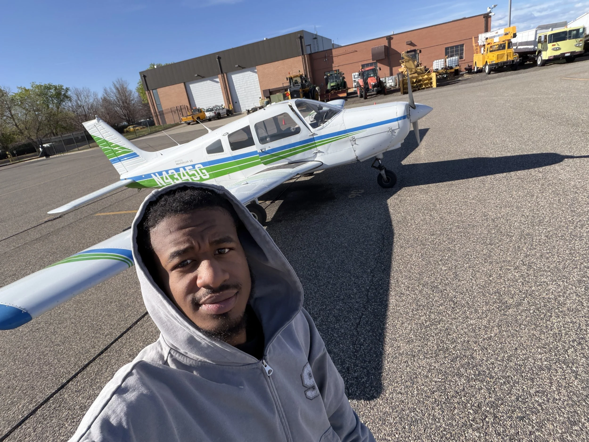 A man taking a selfie in an outdoor parking lot with small aircraft, construction equipment, and building in the background on a clear day.