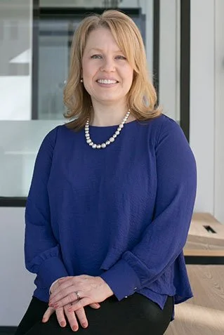 A woman with shoulder-length blonde hair wearing a blue long-sleeve top and a pearl necklace, sitting in a modern office environment.