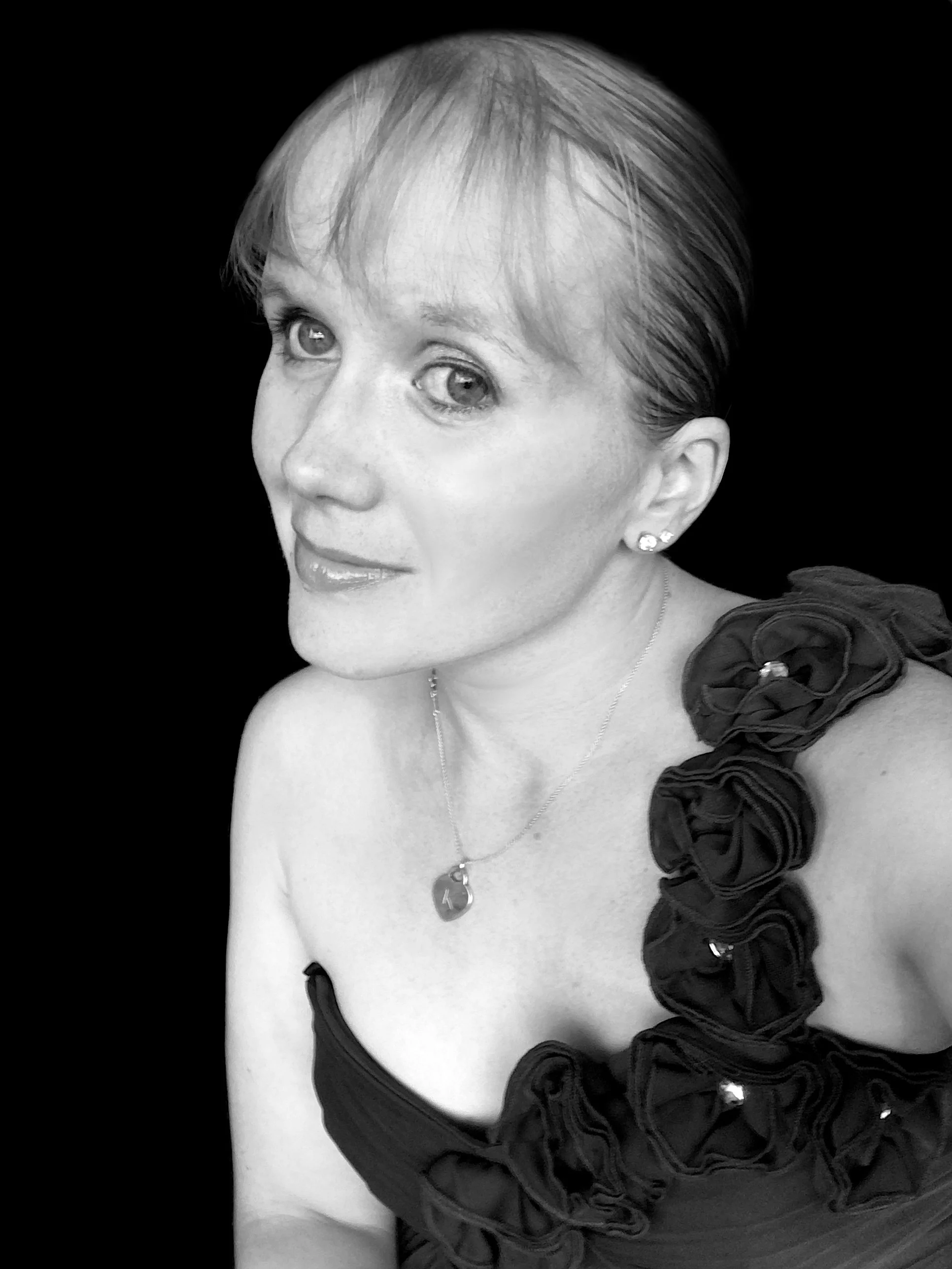 A black and white portrait of a woman with short hair and earrings, wearing a ruffled dress with a large flower on the shoulder, and a pendant necklace with the letter 'K' on it.