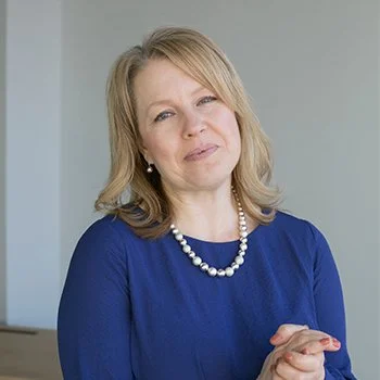 A woman with blonde hair wearing a blue top and pearl necklace, standing indoors against a light-colored wall.
