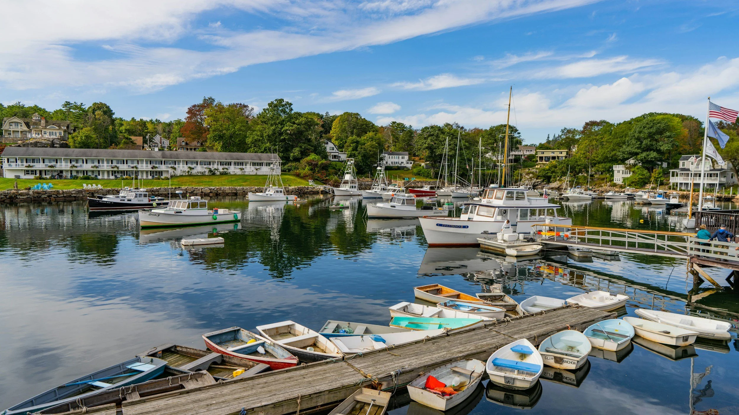 A marina with various boats docked on calm water, surrounded by houses and trees on a clear day.