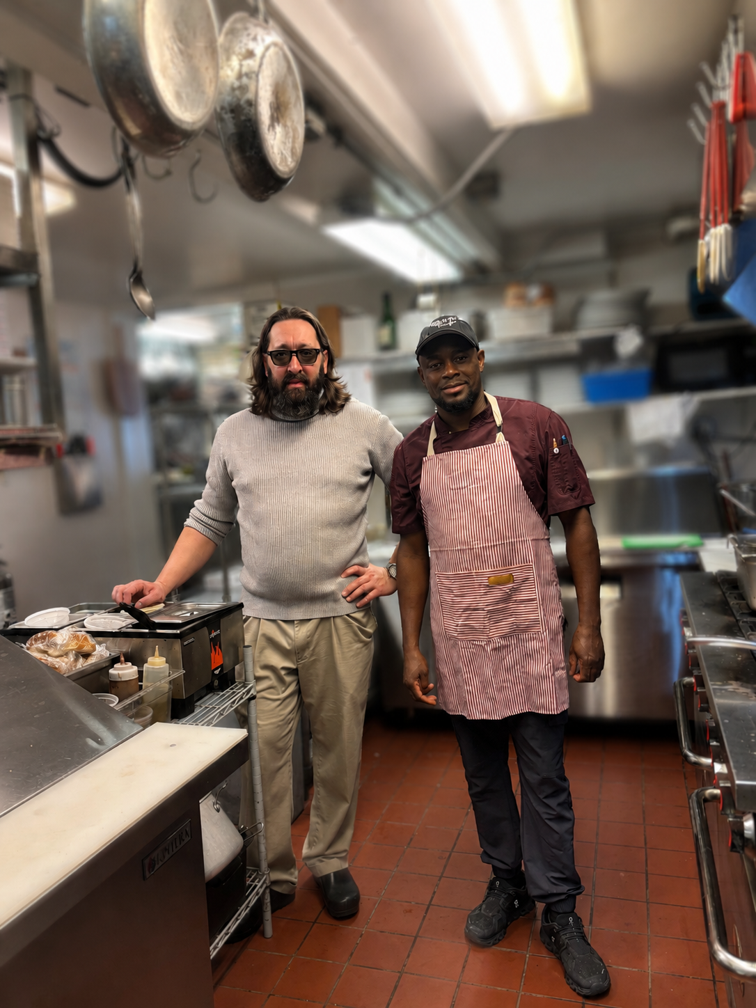 Two men stand in a commercial kitchen. One man has long hair and a beard, wearing glasses, a gray sweater, and khaki pants. The other man has short hair, wearing a chef's hat, a maroon chef's coat, and an apron. There are kitchen utensils and appliances visible around them.