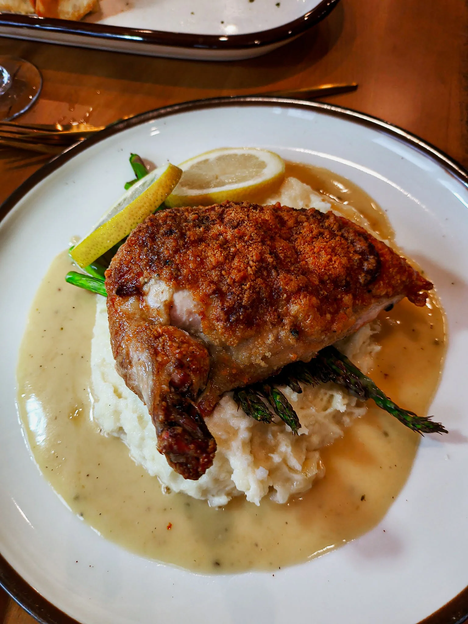 A plate with a serving of mashed potatoes topped with a roasted chicken leg, garnished with grilled asparagus, lemon slices, and a sprig of herbs, all in gravy.