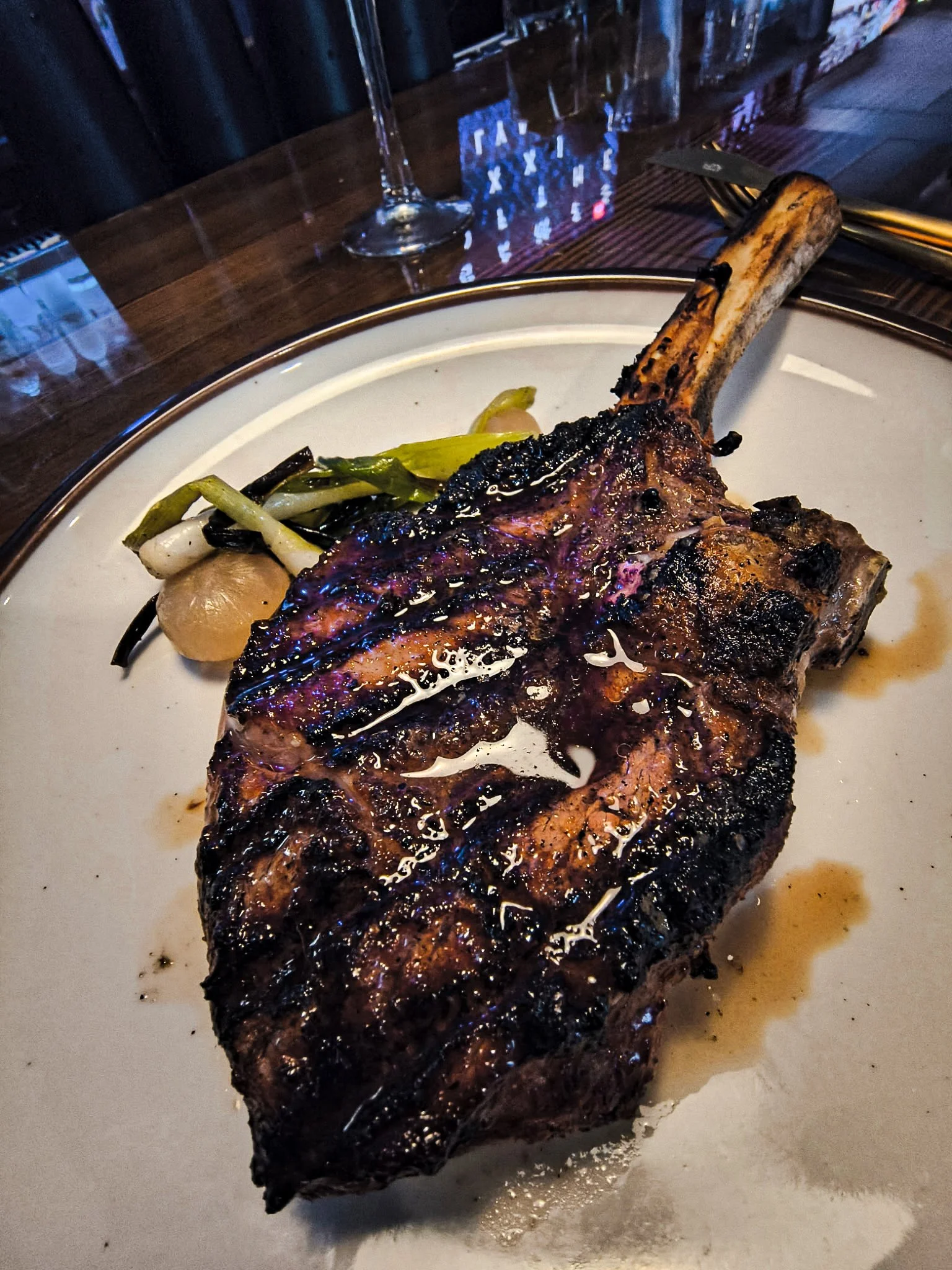 A grilled steak with a bone, served on a white plate with some grilled vegetables and potatoes.