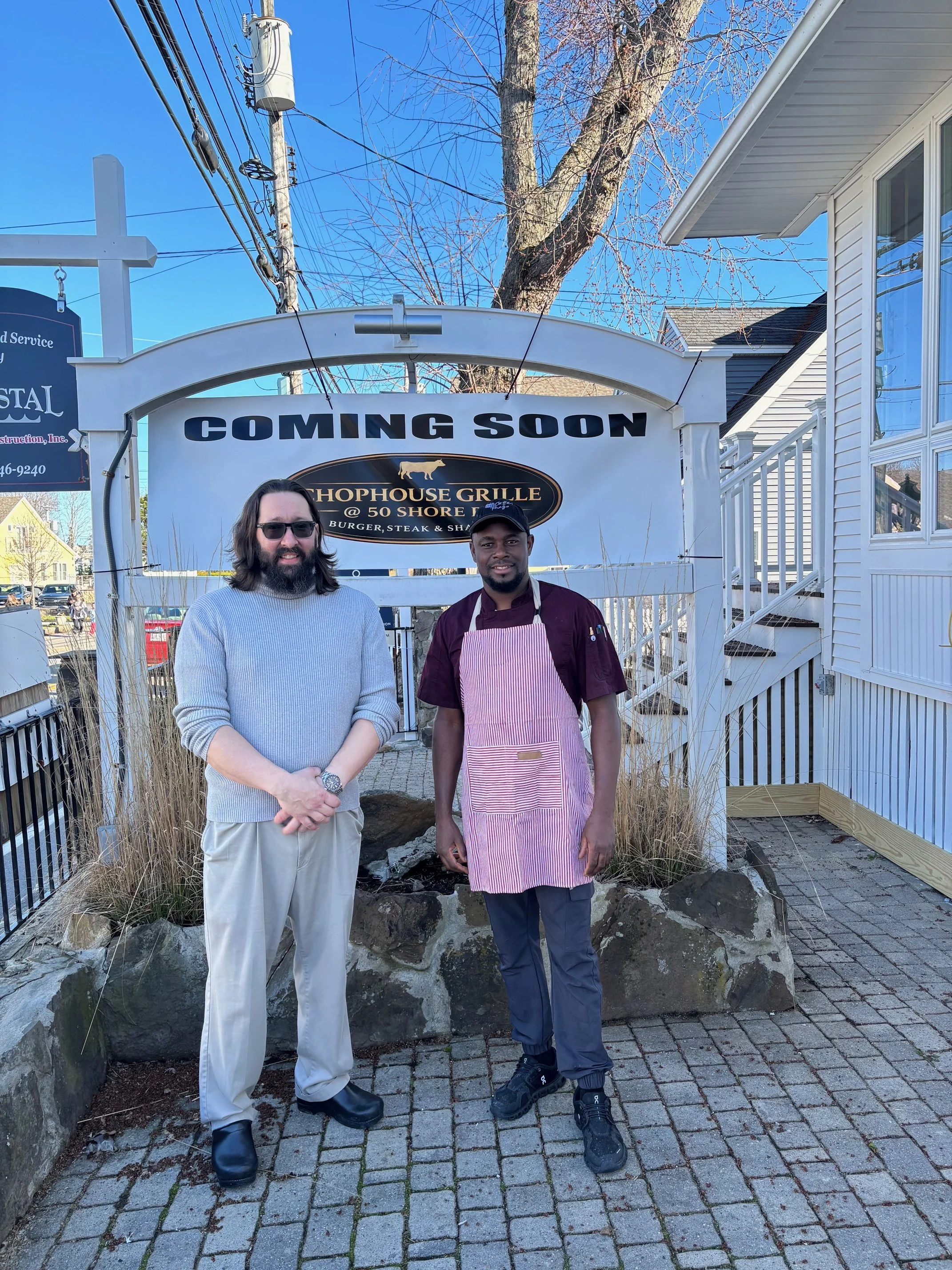 Two men standing outdoors in front of a sign that reads "COMING SOON" for the Shohouse Grille. One man has long hair and a beard, wearing sunglasses, a light gray sweater, beige pants, and black boots. The other man has short hair, wearing a maroon shirt, a pink and white striped apron, and black shoes. There is a clear blue sky, leafless trees, and residential houses in the background.