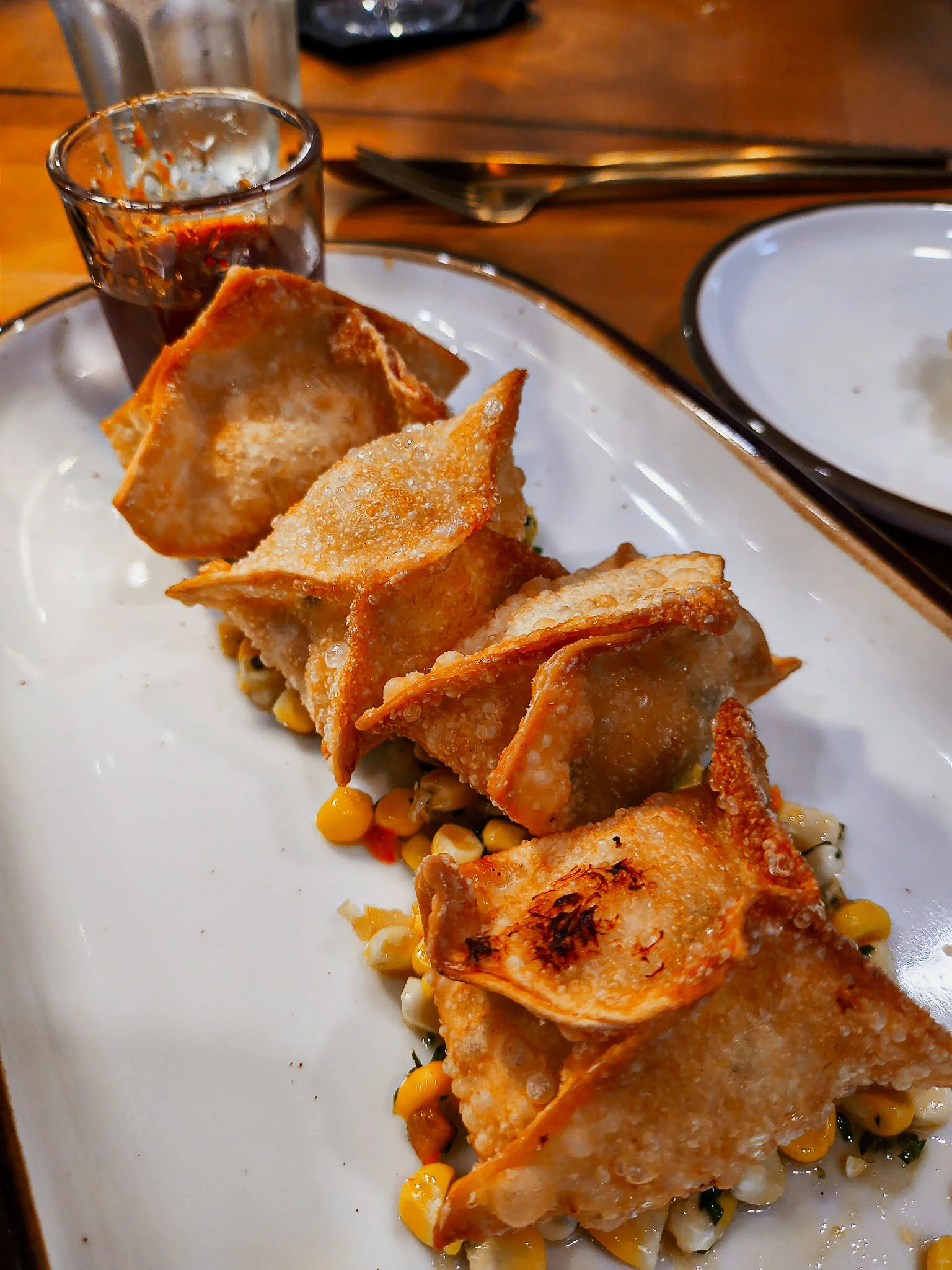 Close-up of a plate with fried dumplings on top of a bed of mixed vegetables, including corn and greens, with a small glass of soy sauce and a dipping bowl visible in the background.