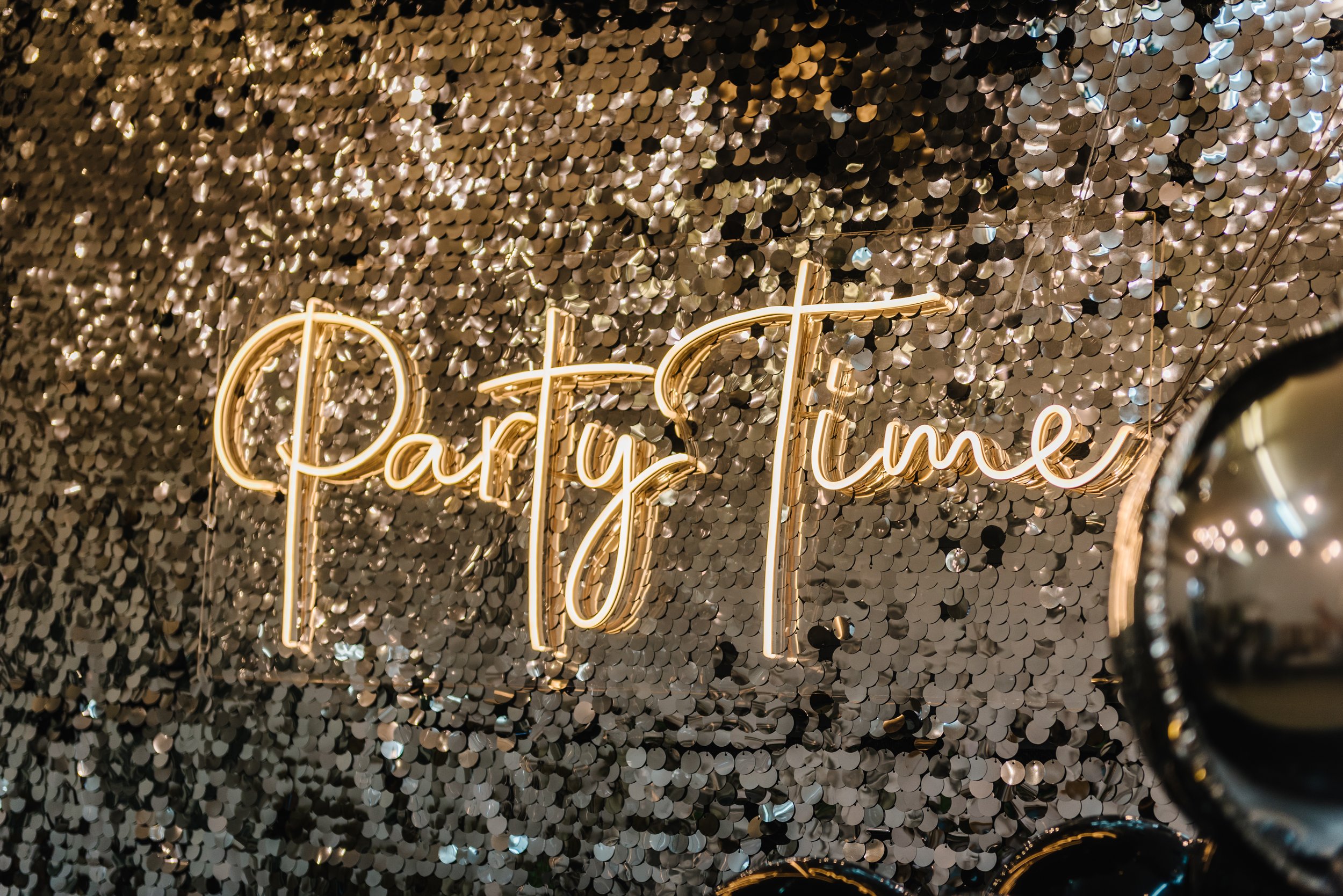 Leuchtendes Neon-Schild mit dem Wort 'Partytime' vor einer Wand aus silbernen Pailletten.