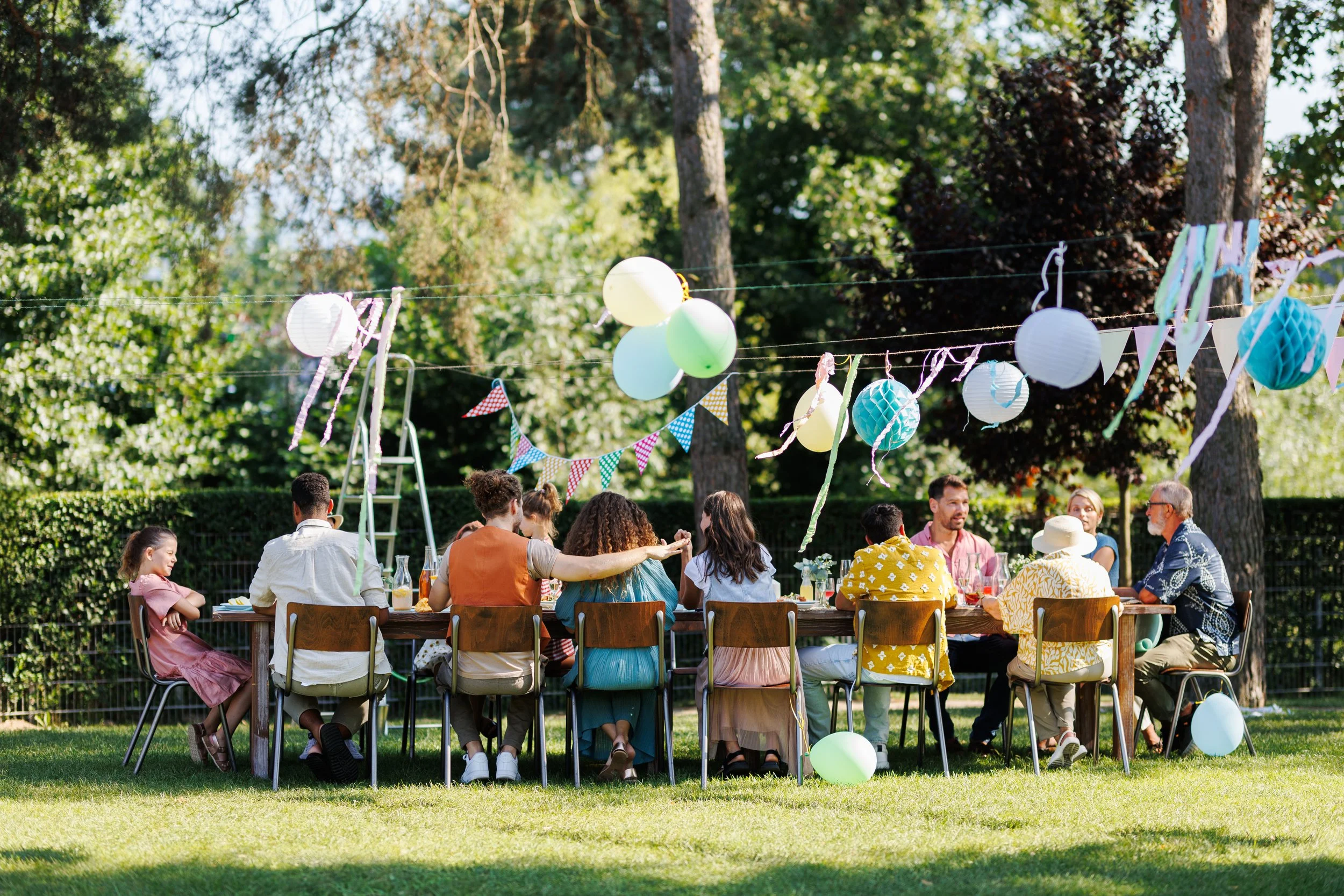 Gruppe von Menschen bei einer Gartenparty im Freien, dekoriert mit bunten Papierlaternen, Ballons und Wimpeln, umgeben von Bäumen.