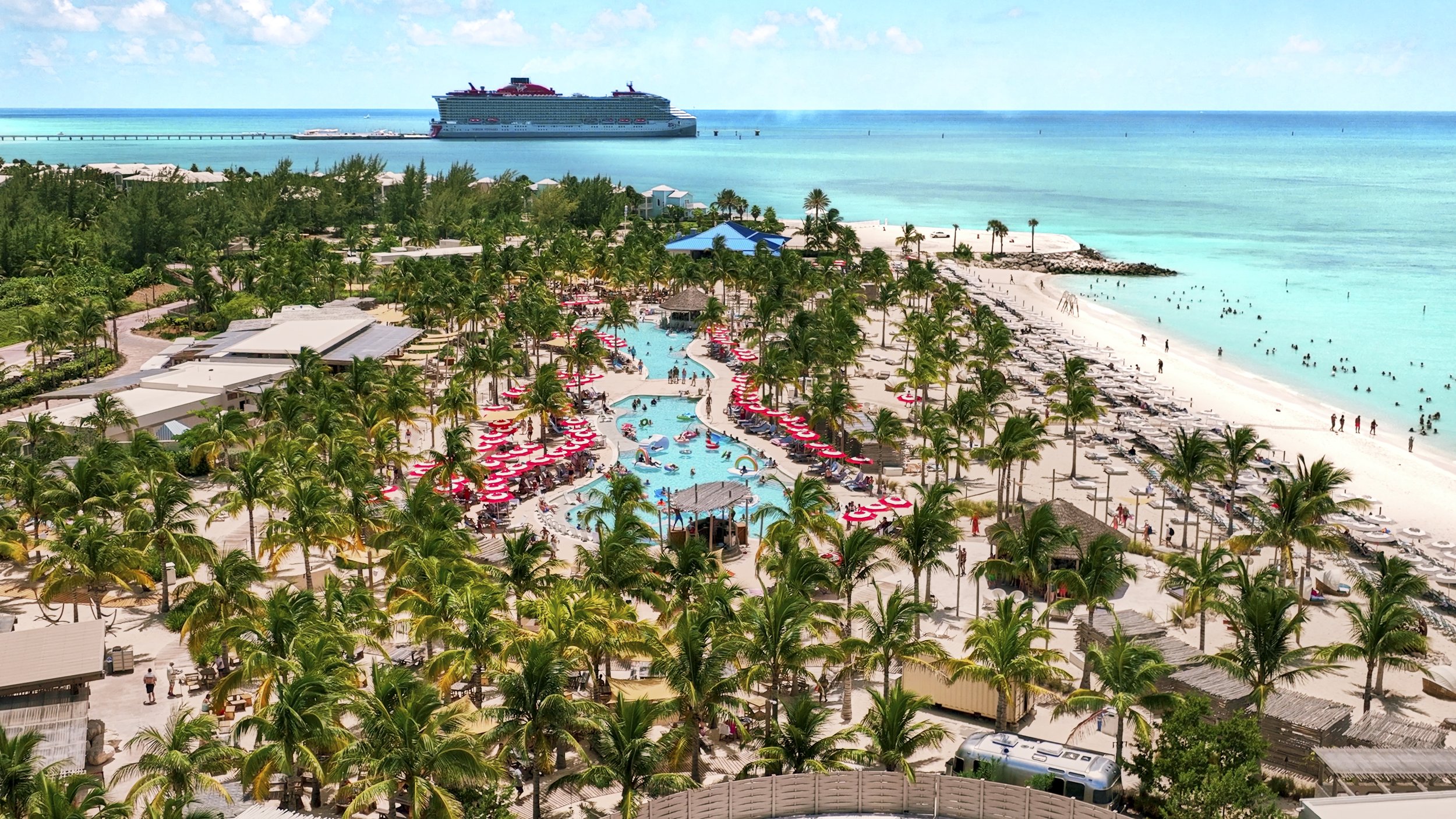 Aerial view of a tropical beach resort with a large cruise ship docked nearby in turquoise ocean. The resort has a lazy river, swimming pools, palm trees, umbrellas, and lounge chairs, with visitors enjoying the water and relaxing.