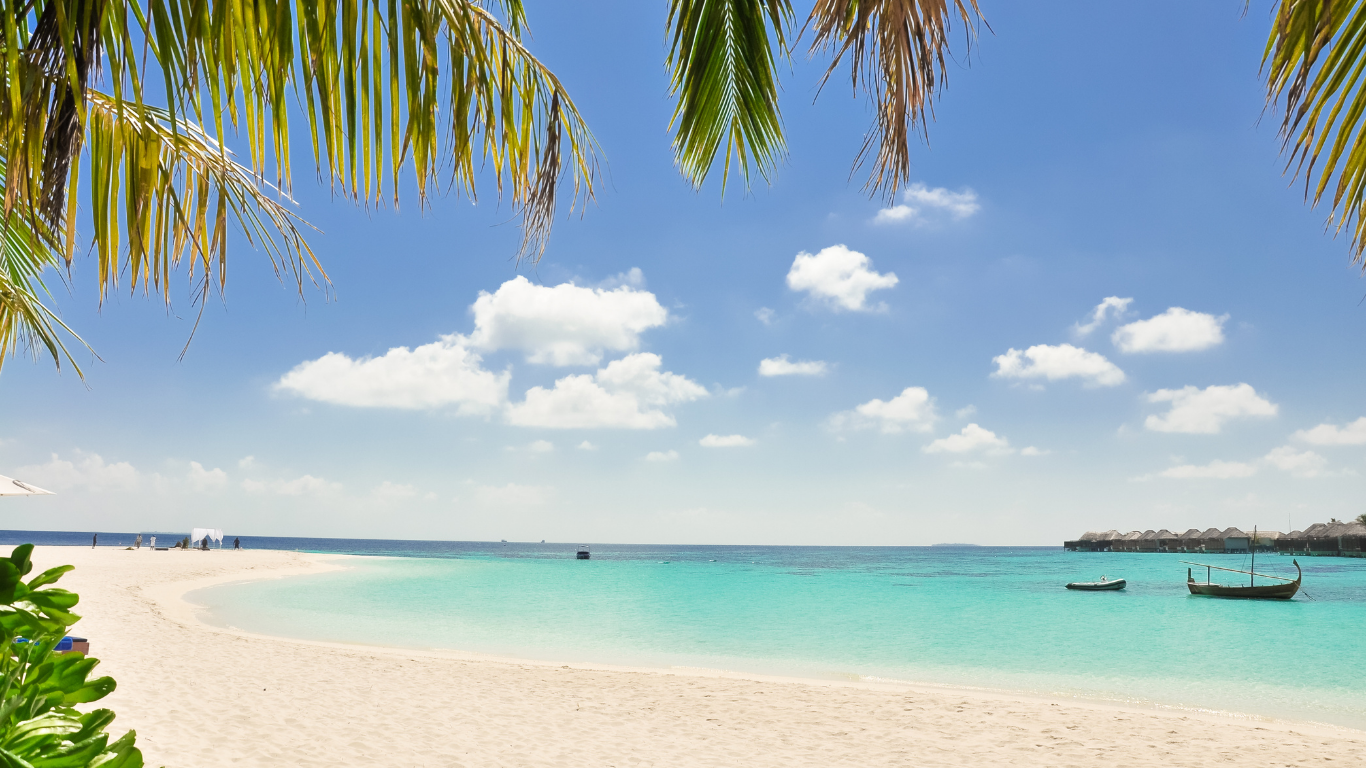 Tropical beach with white sand, clear turquoise water, palm tree leaves overhead, and small boats floating in the water with overwater bungalows in the background under a blue sky with fluffy white clouds.