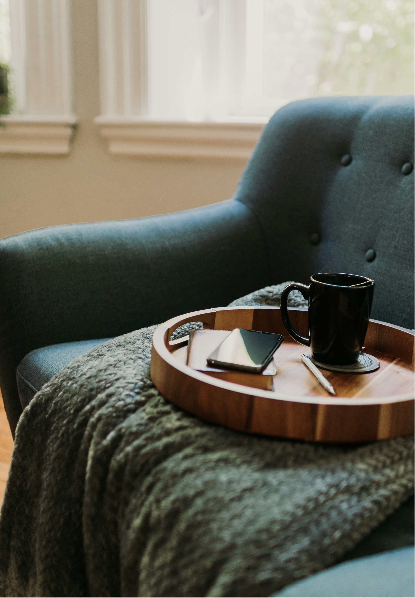 A cozy living room scene with a round wooden tray on a textured blanket, holding a black mug, a smartphone, a notebook, and a pen, next to a blue upholstered armchair with button tufting, near a window with white curtains.